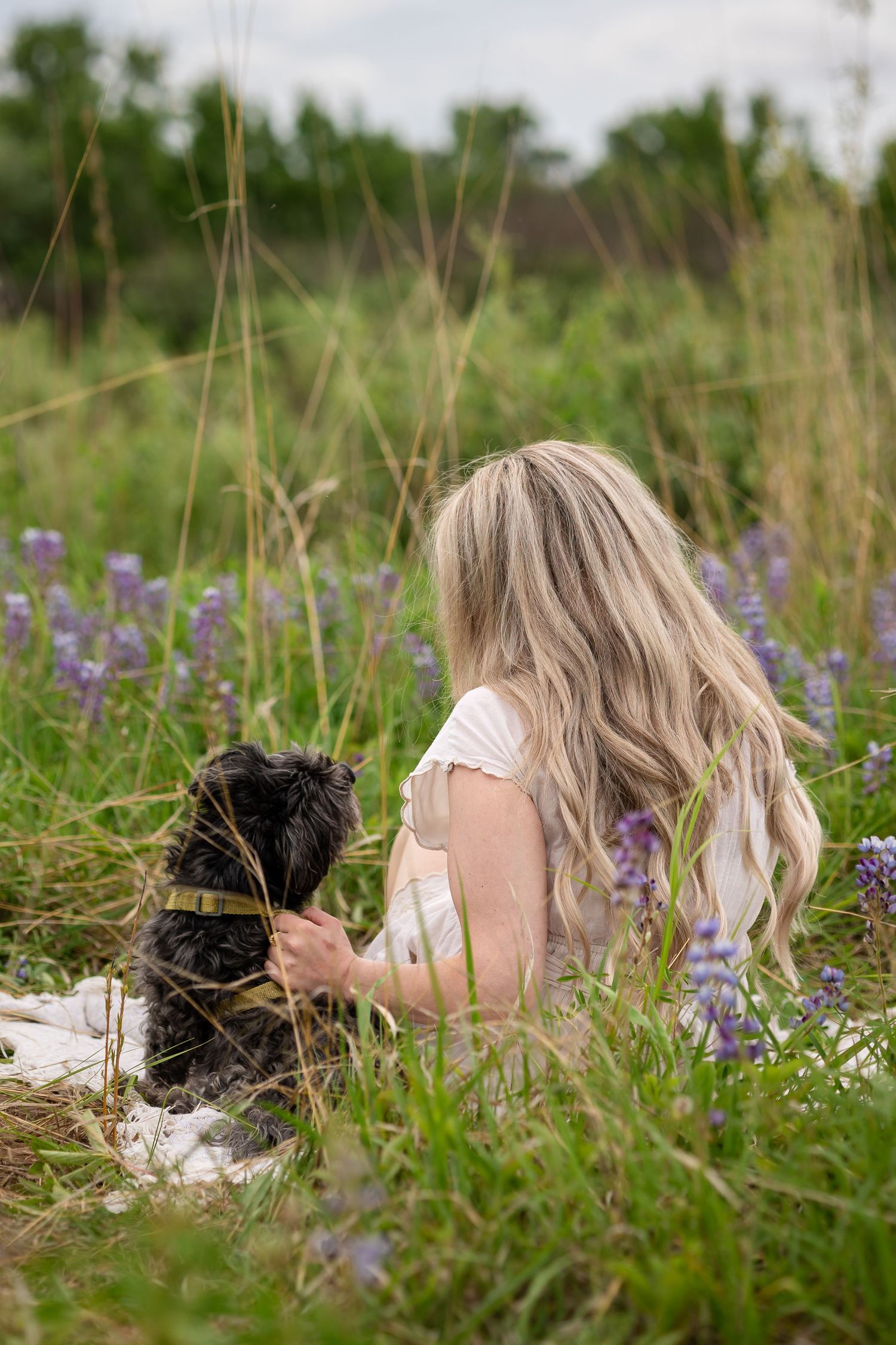 Blonde woman in white dress sitting in a field of purple wildflowers with her small black dog on a sunny day.