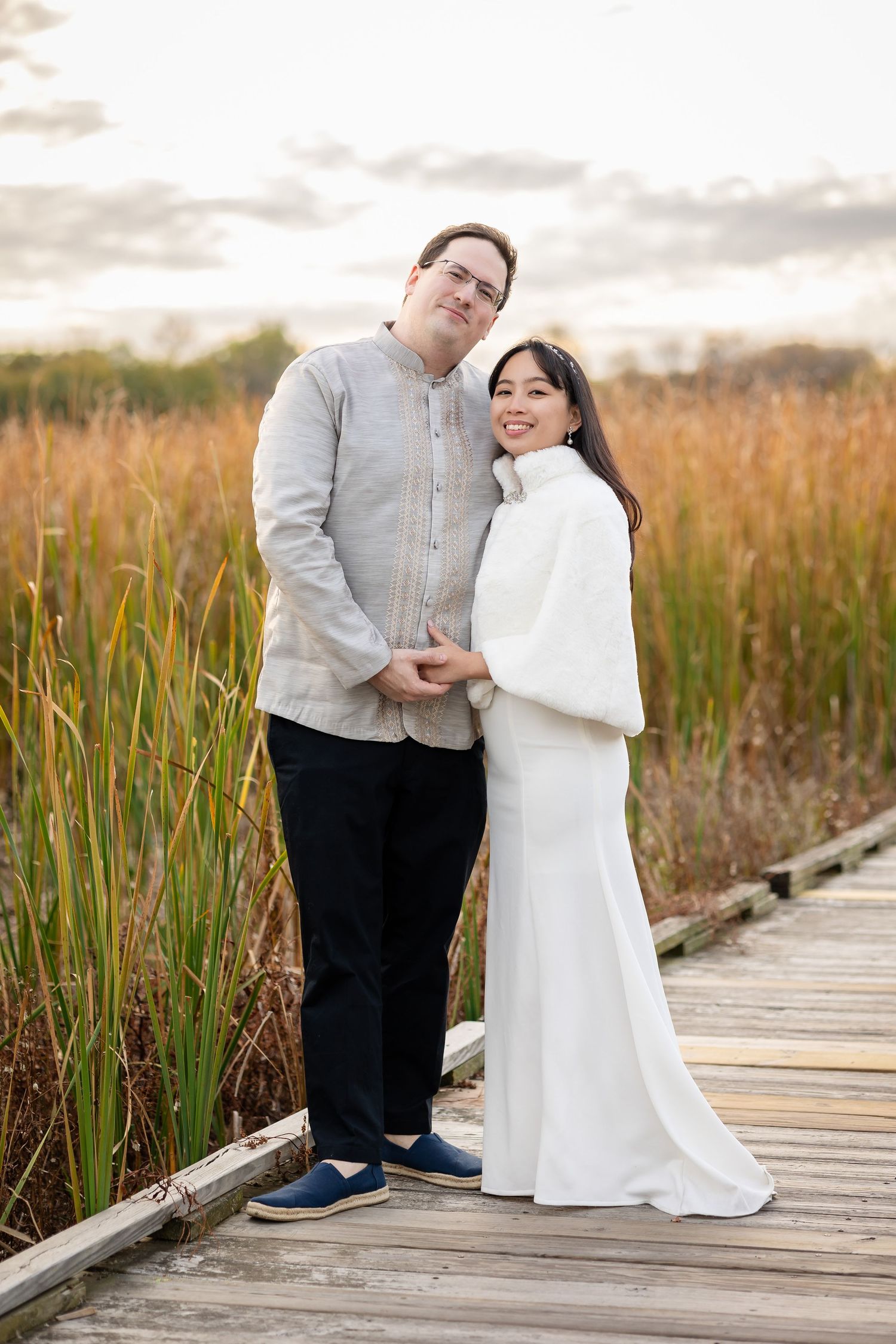 A couple poses on a wooden boardwalk surrounded by tall autumn grasses at sunset.