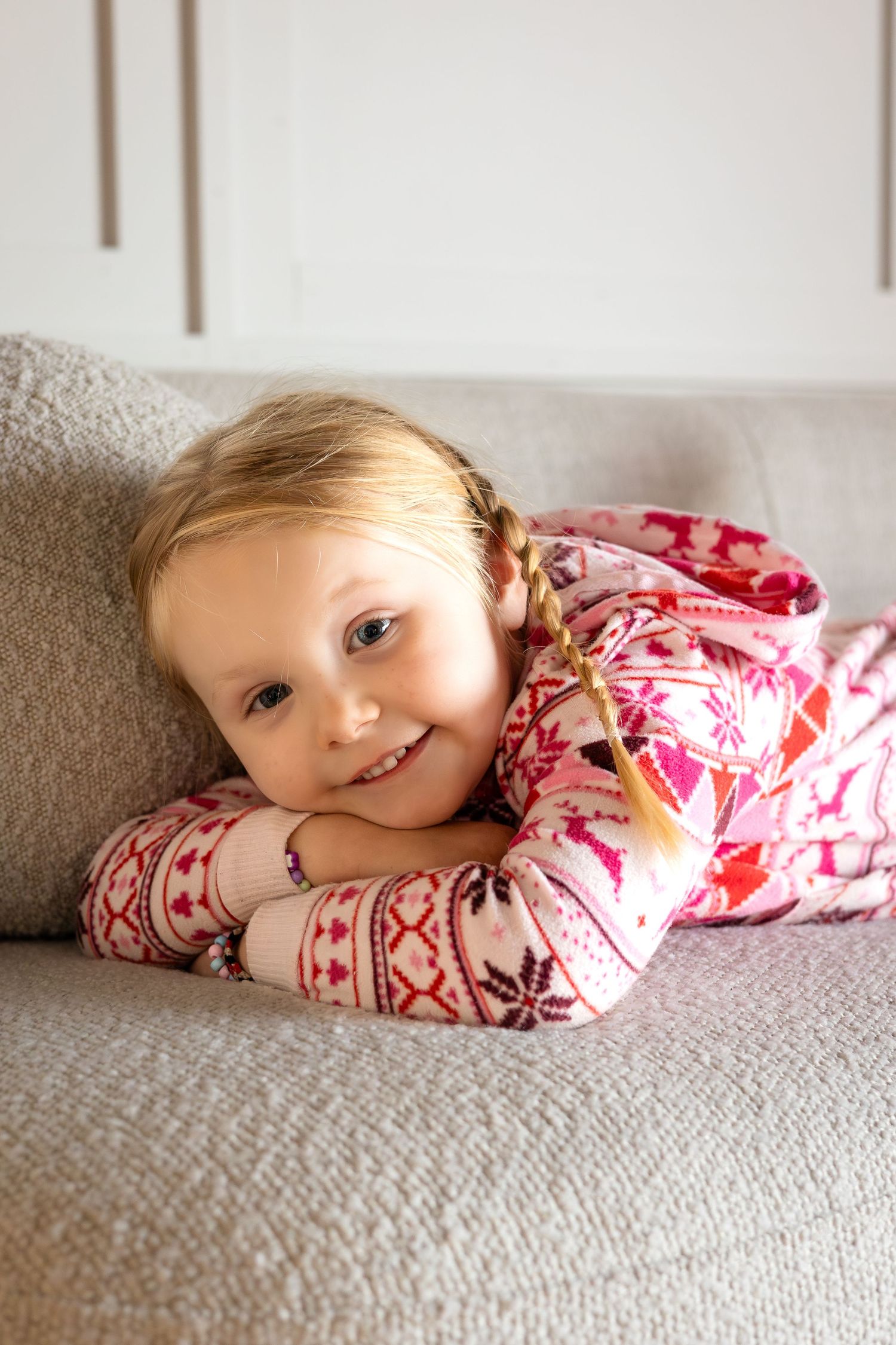 Child in pink patterned Christmas pajamas lying on gray couch with sweet expression.