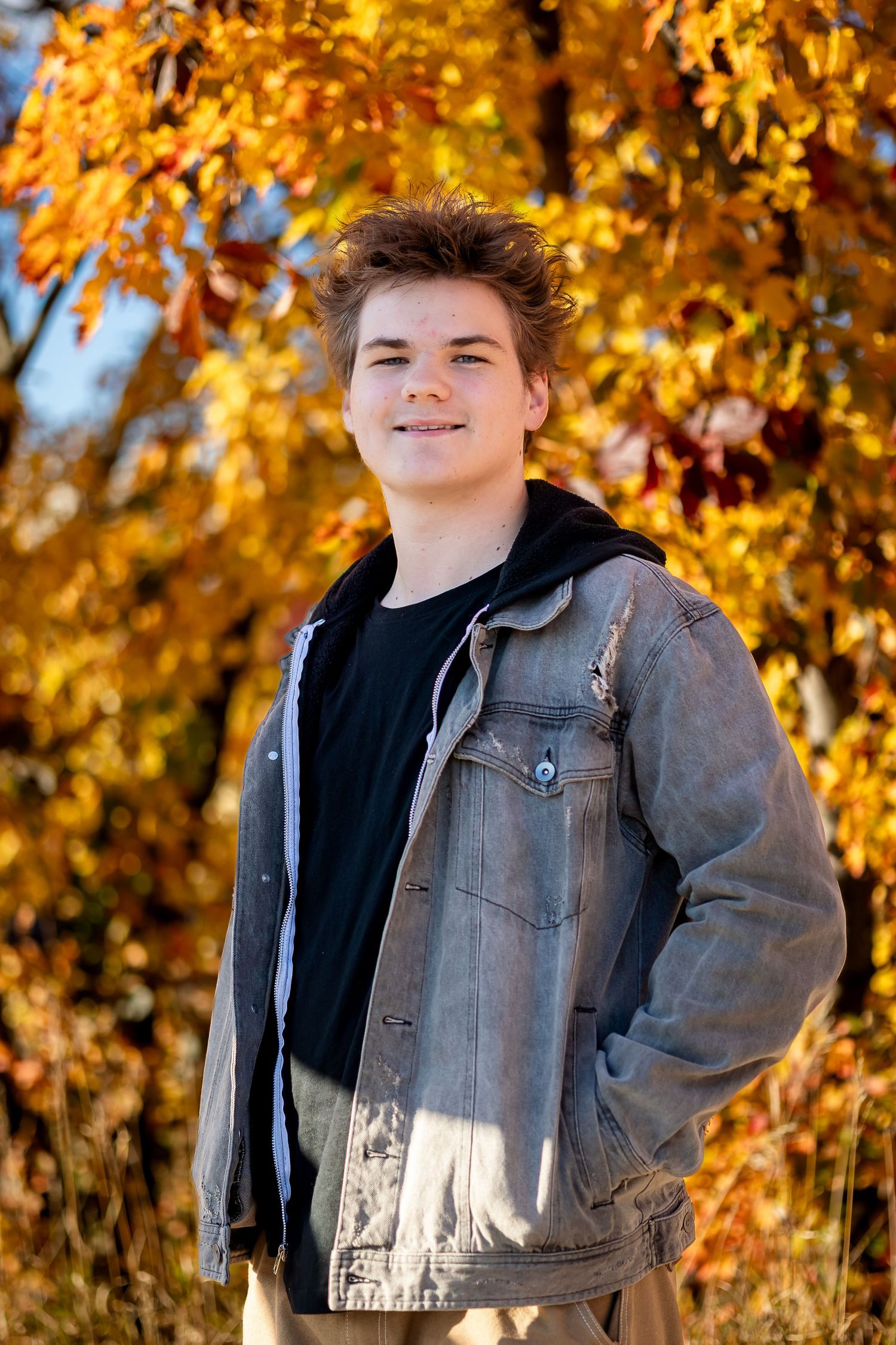 Person wearing a denim jacket poses against vibrant autumn foliage with golden yellow leaves in background.