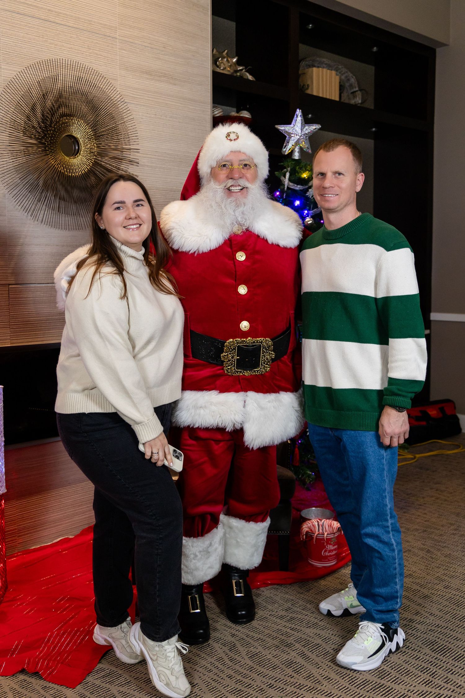 A Christmas gathering with Santa in his bright red suit standing on a red carpet near a decorated tree.