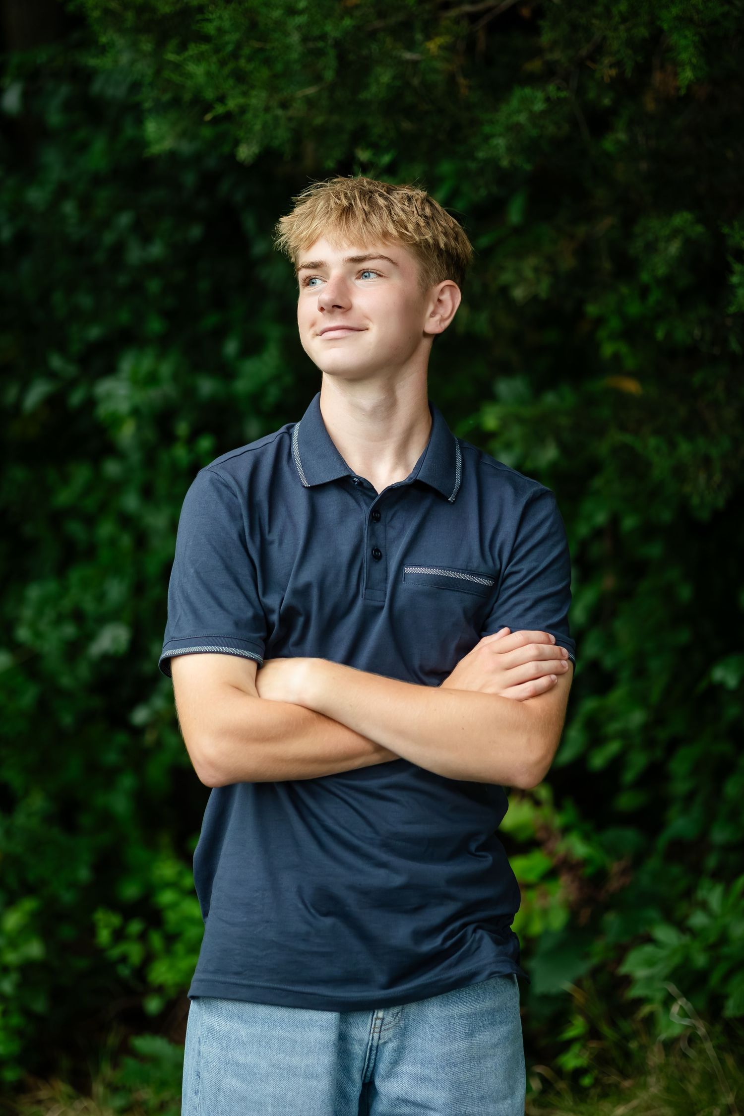 Person in navy polo shirt poses with crossed arms against lush green foliage.