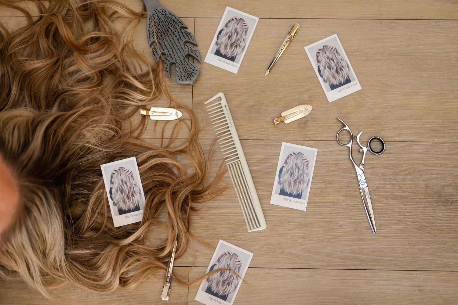 Hair styling tools and Polaroid photos arranged on a light wooden surface with blonde hair extensions visible.