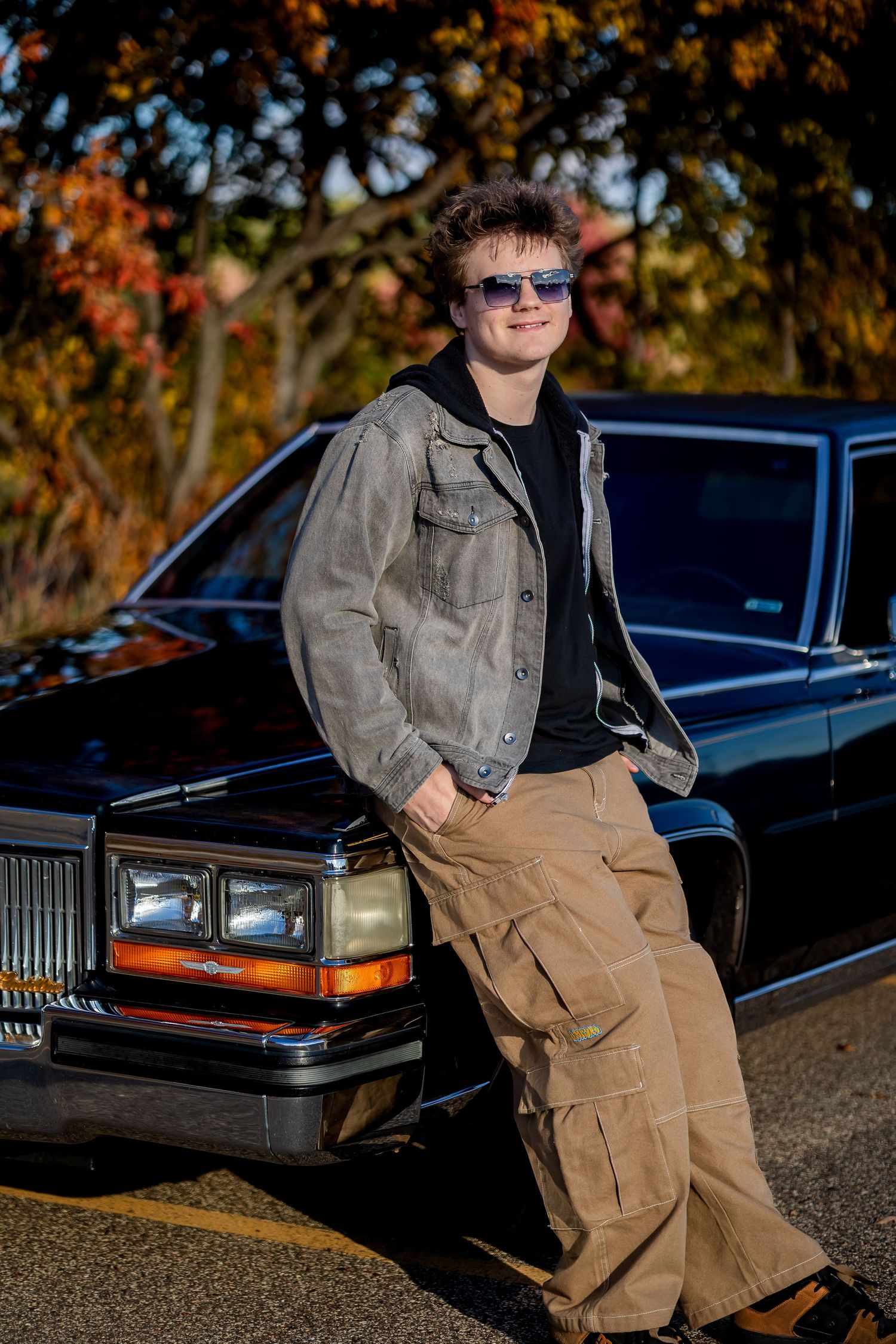 A vintage Cadillac sedan parked outdoors during autumn with someone posing near it in casual attire.