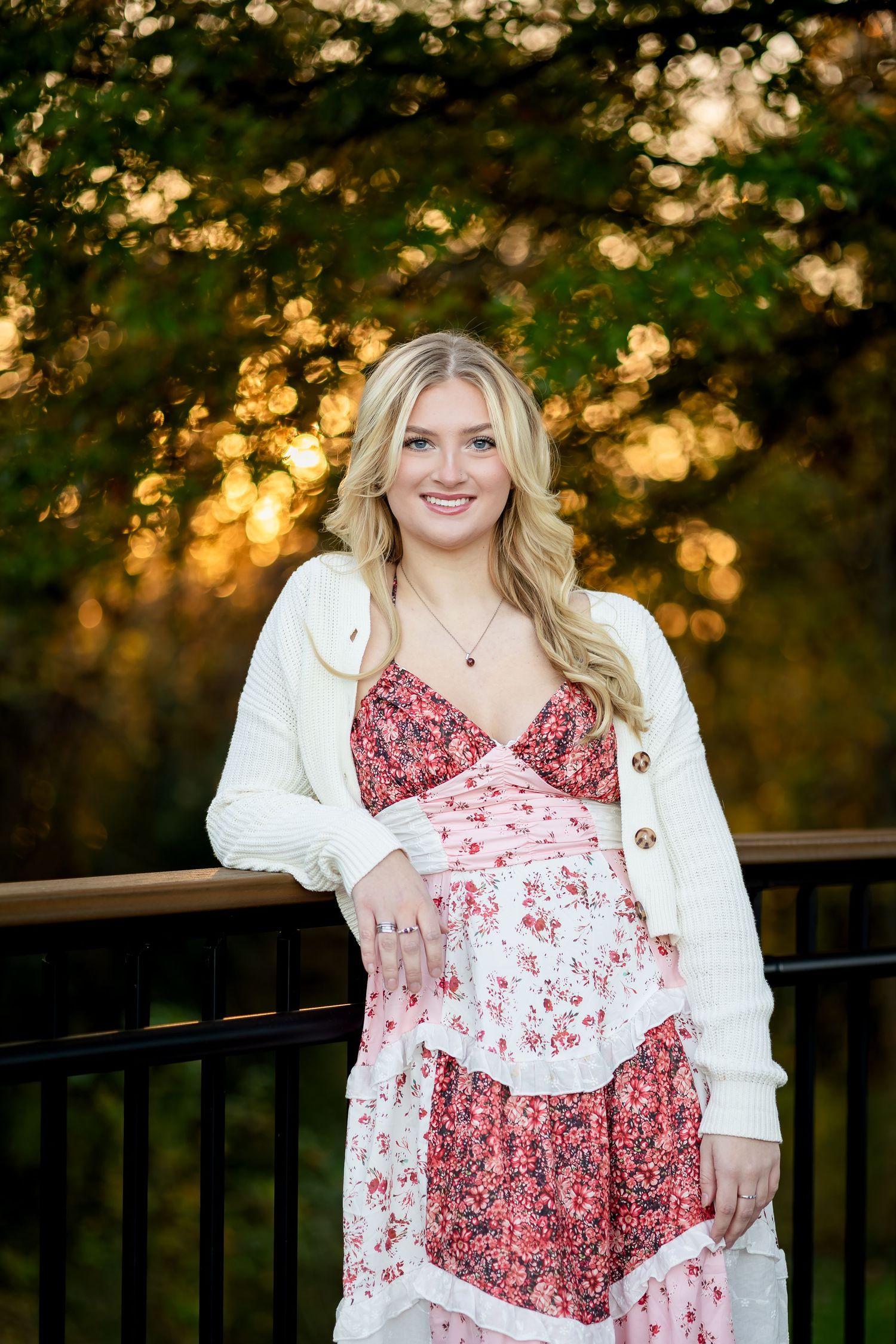 A woman in a red and white patterned dress poses on a wooden deck during sunset.