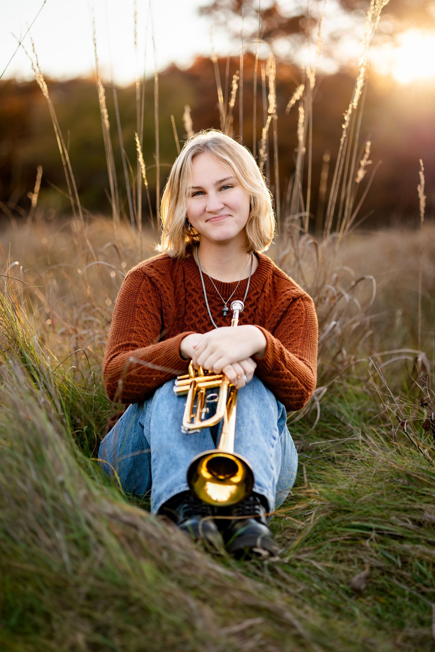 A person sits in tall grass at sunset holding a brass musical instrument.