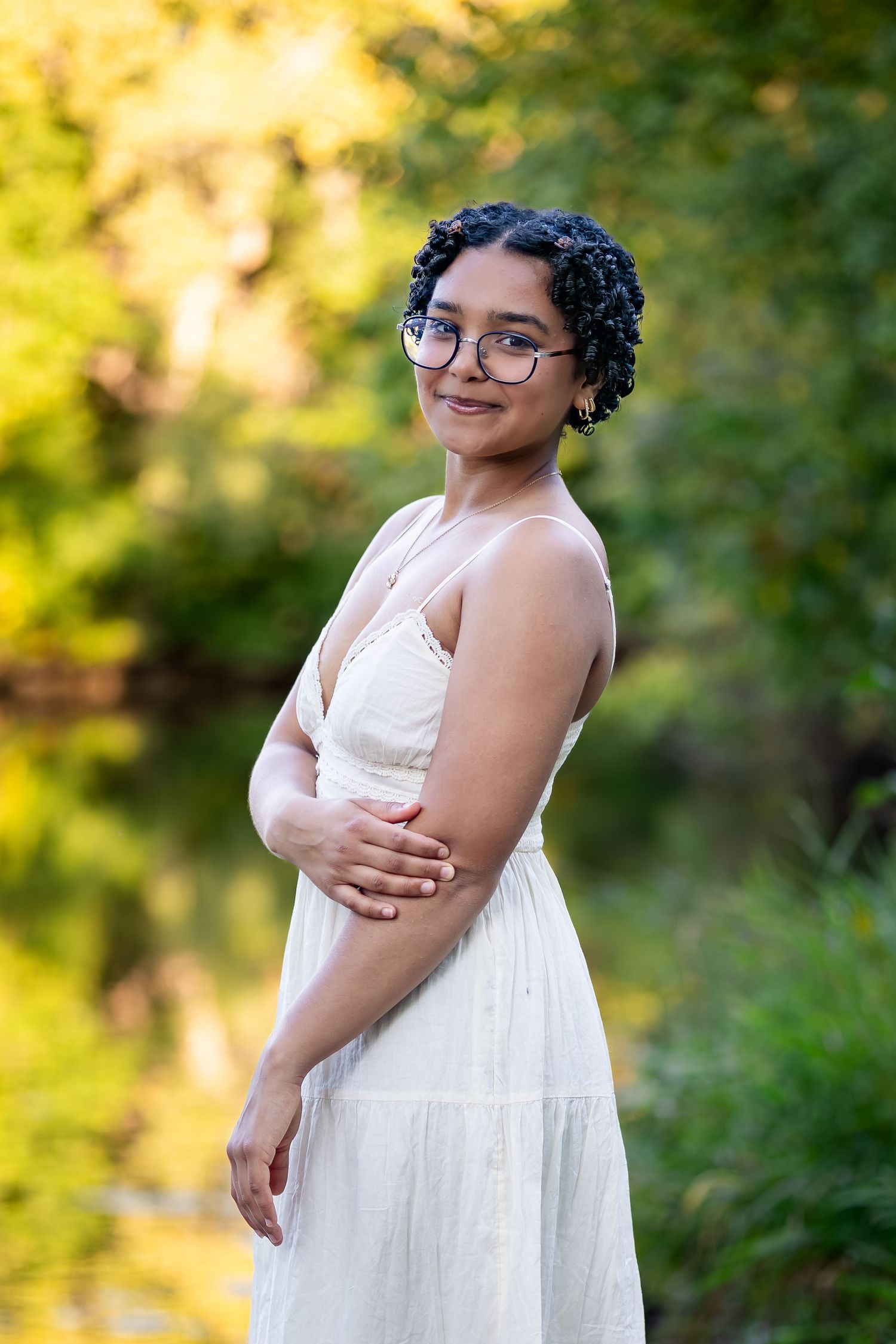 A person in a flowing white dress poses outdoors in a garden setting with warm golden sunlight filtering through trees.