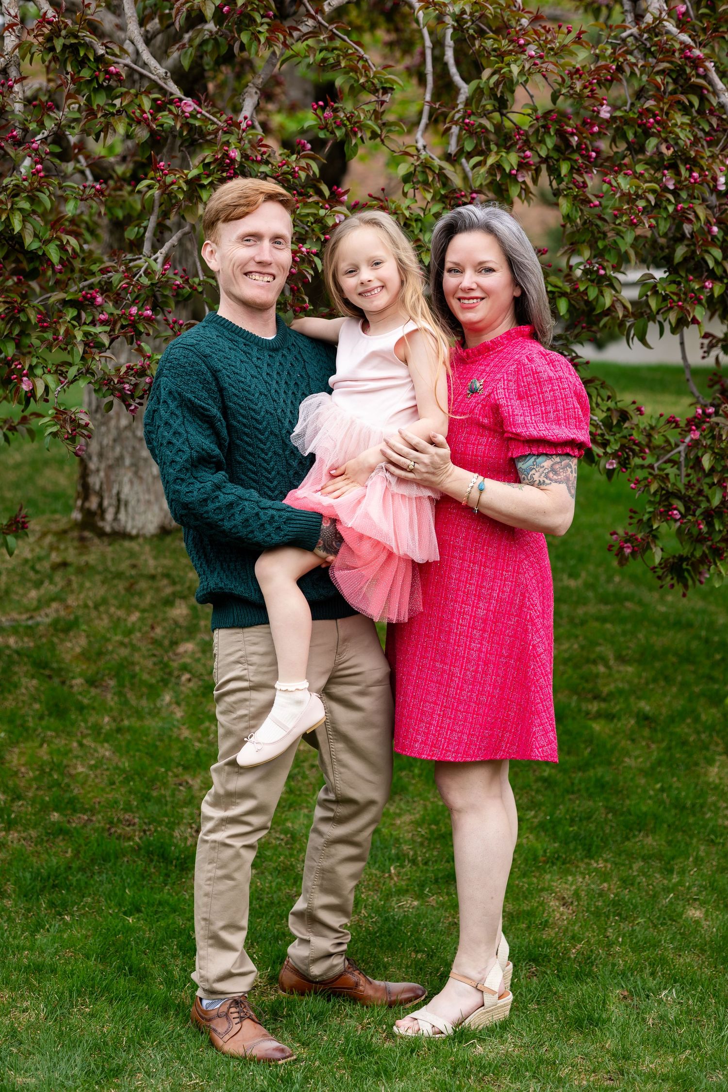 Happy family of three posing outdoors under blooming cherry tree, parents holding young daughter in pink tutu.