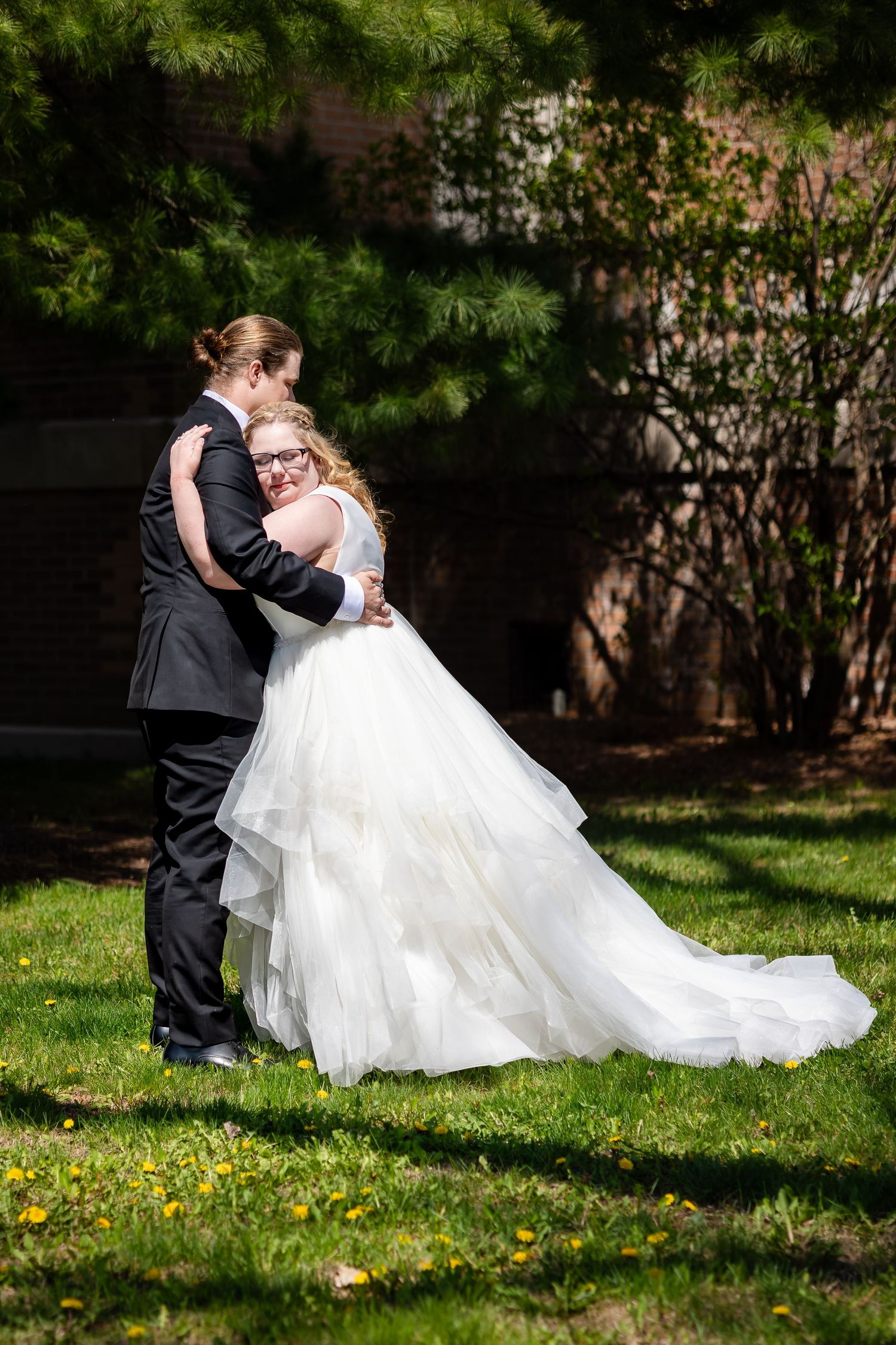 Groom in black suit embracing bride in white layered gown outdoors on green lawn.