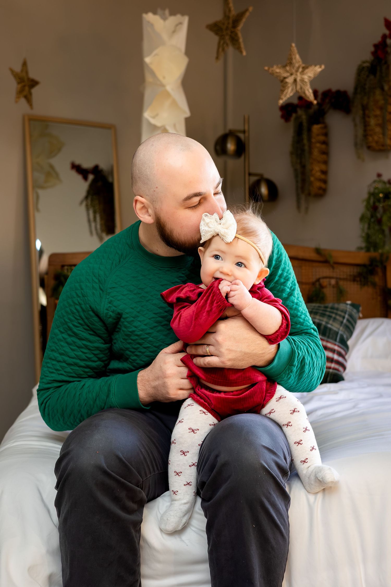 Bald father in green sweater kissing baby girl in red dress with white bow headband on bed with Christmas decorations.