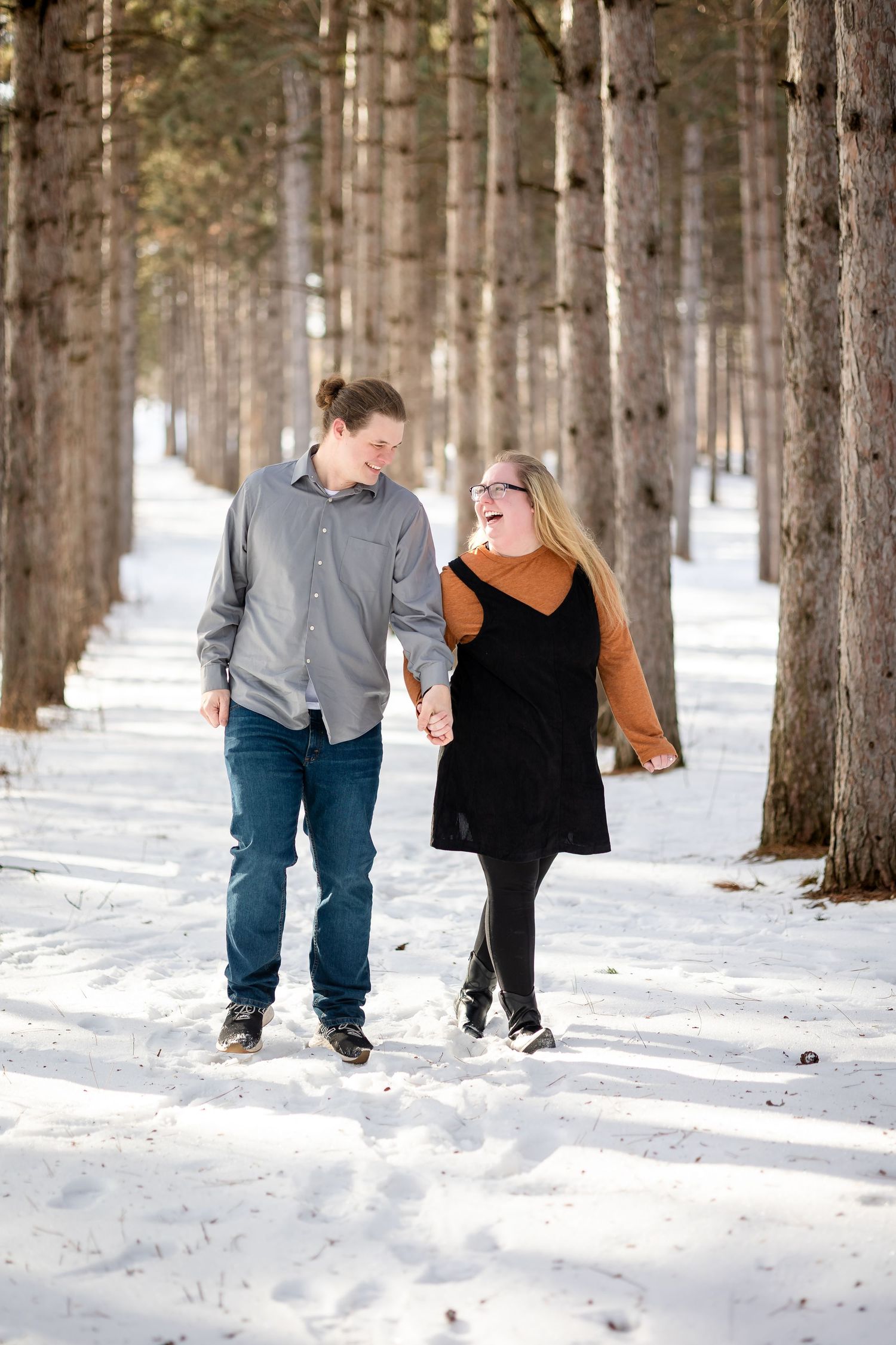 Couple holding hands and laughing while walking through a snowy pine tree lined path in winter.