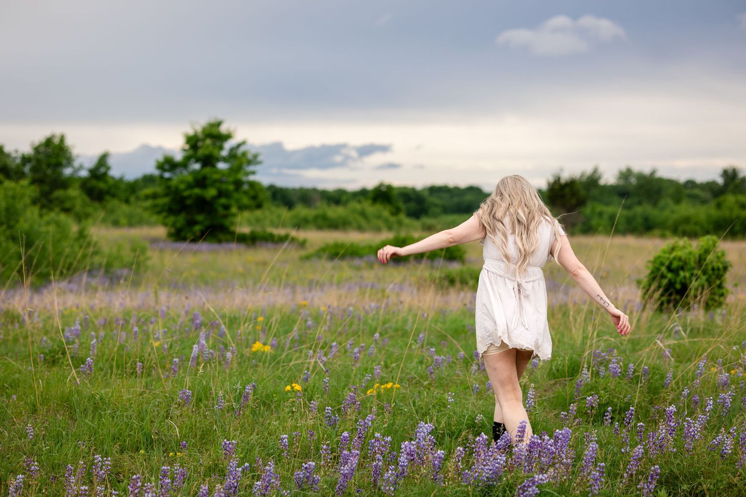 Blonde woman in white dress twirling in a field of purple wildflowers under cloudy skies.