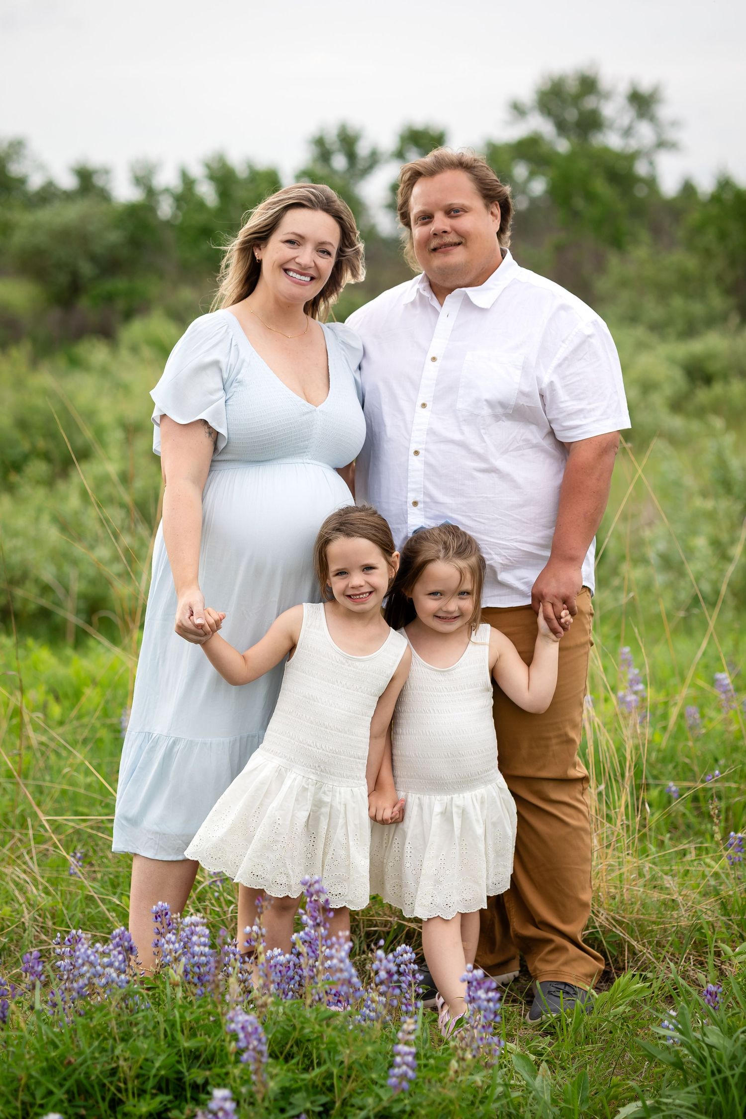 Pregnant woman in blue dress poses with husband and two young daughters in white dresses among purple wildflowers.