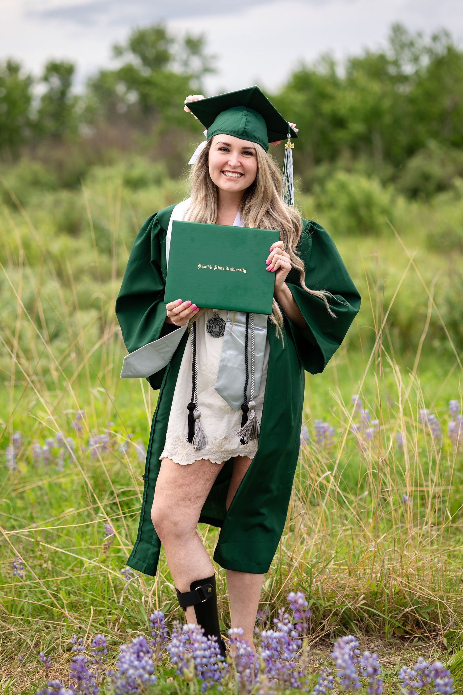 Smiling graduate in green cap and gown holds diploma in a wildflower field, celebrating her Portland State University degree.