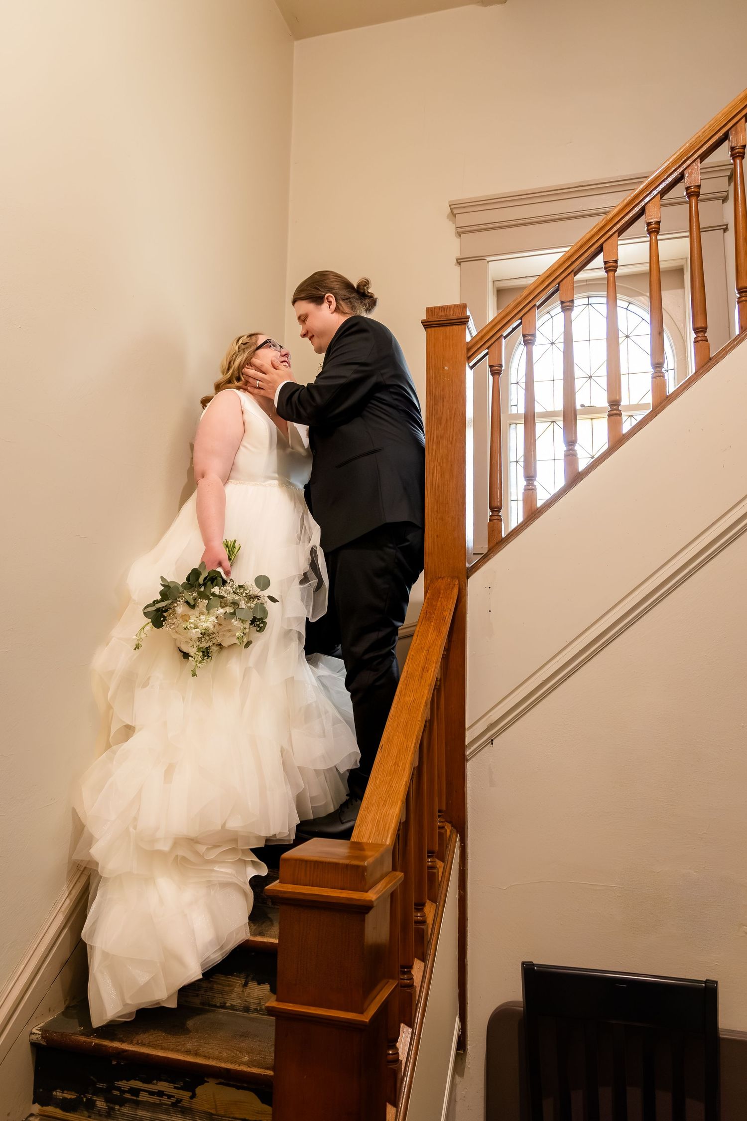 Bride and groom share tender moment on wooden staircase with stained glass window.