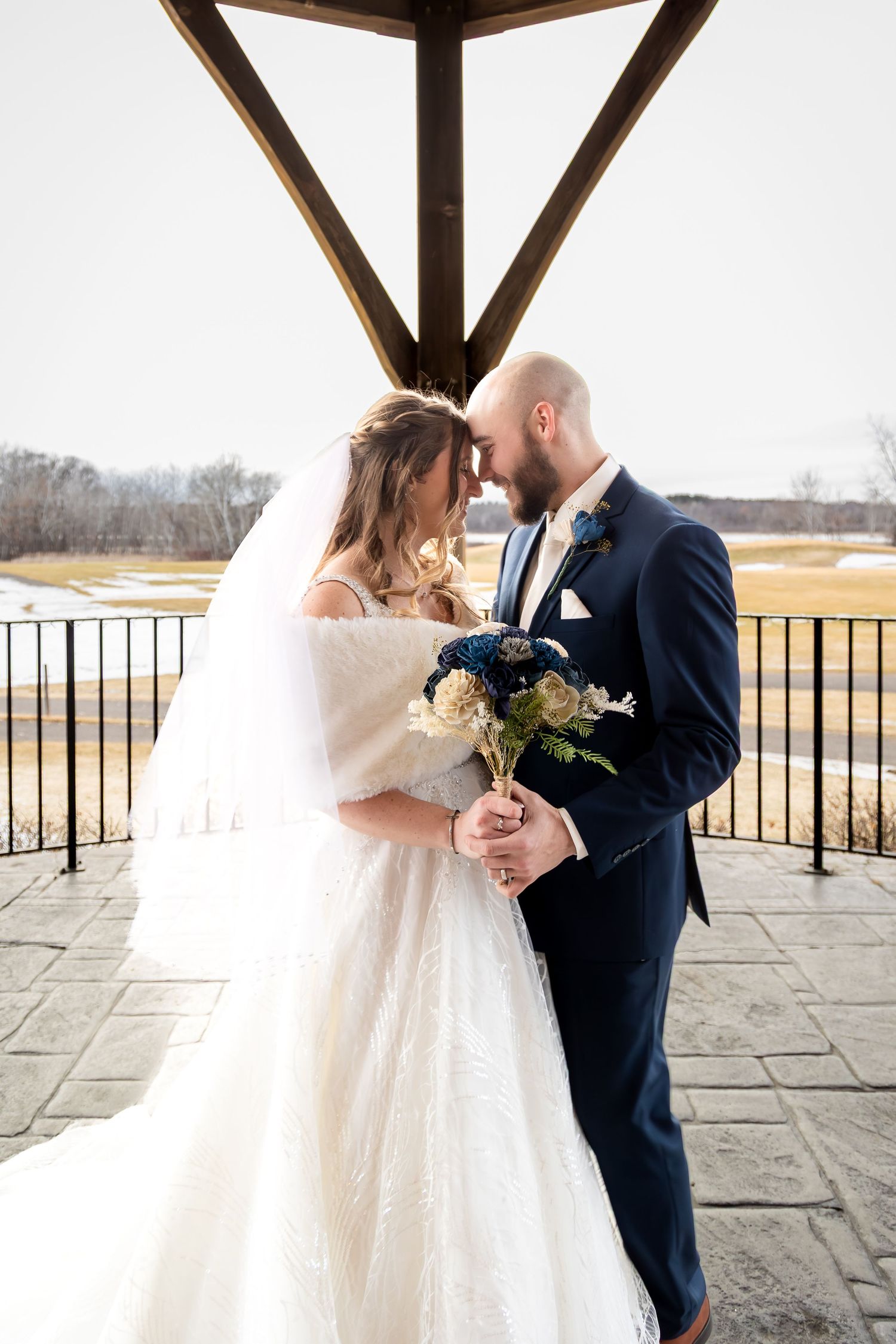 Bride and groom share intimate forehead touch holding blue and white bouquet outdoors.