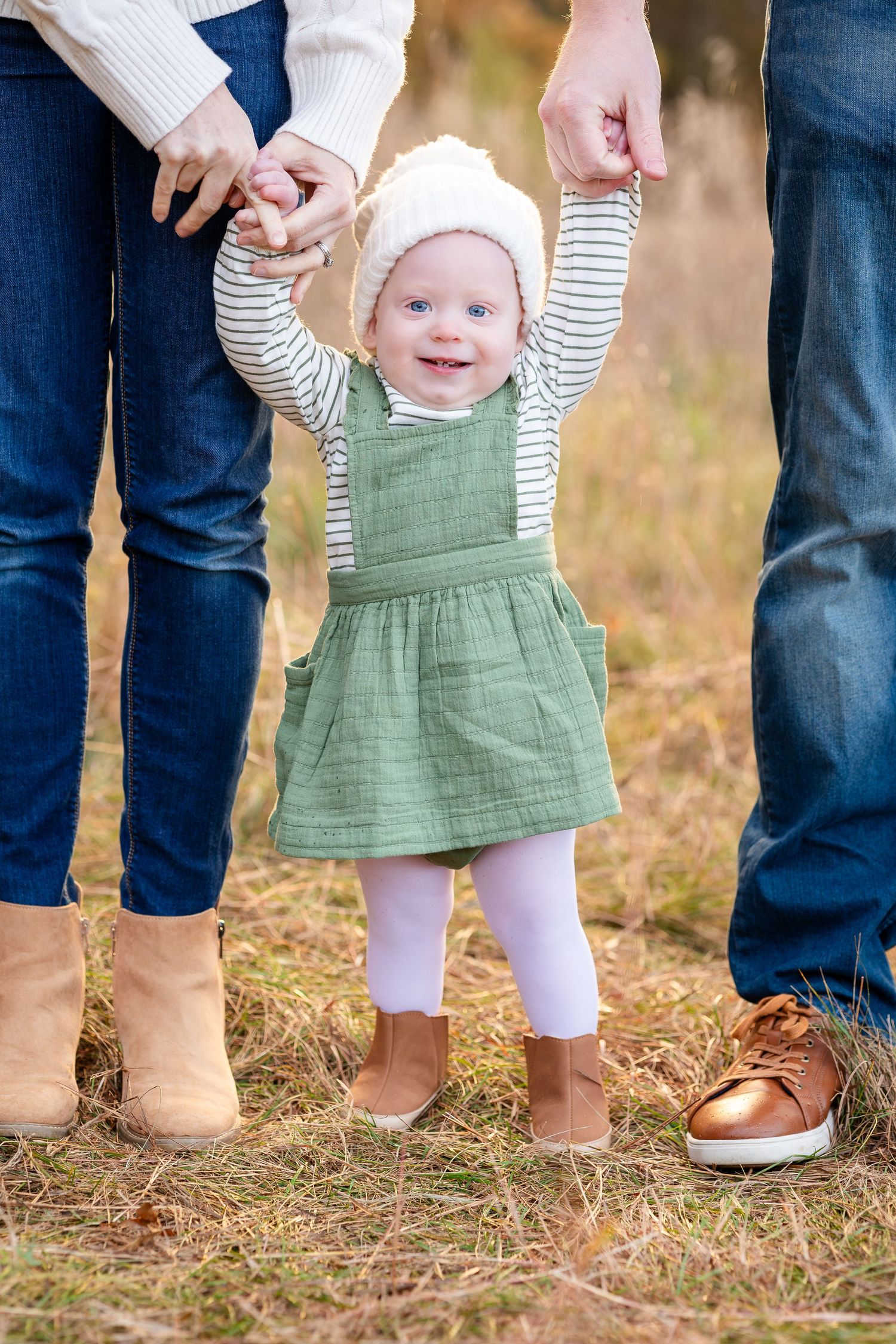 A baby in a green dress takes first steps while holding parents' hands outdoors.