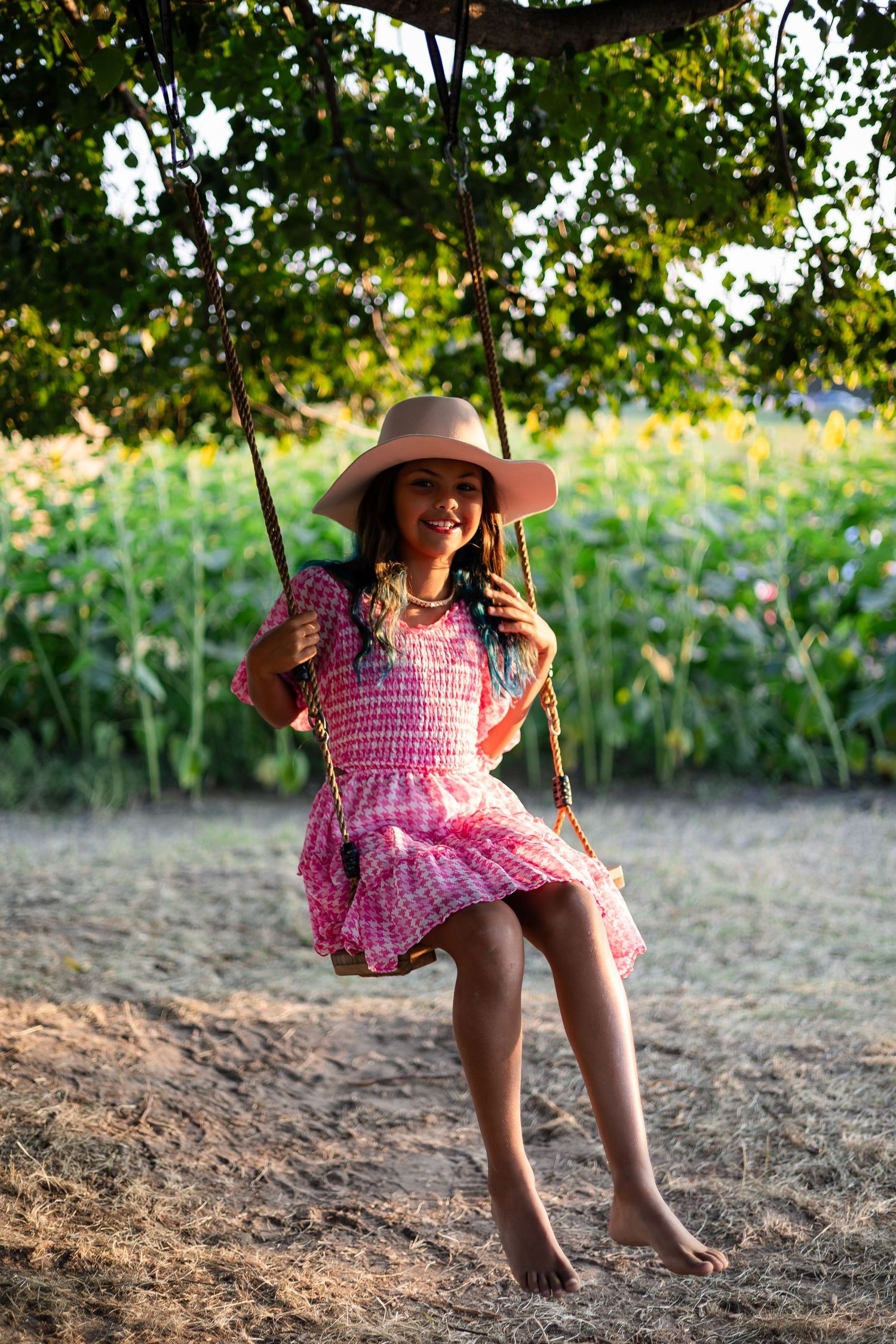 A child in a pink dress swings happily beneath shady trees near a sunflower field.