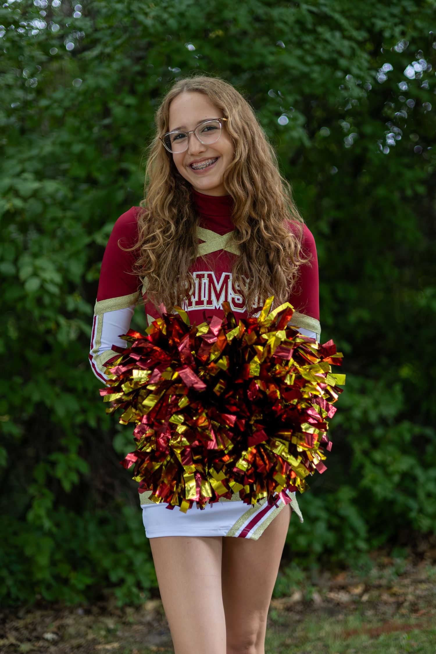 A cheerleader in a red and white uniform holds metallic pom poms while posing outdoors against green foliage.