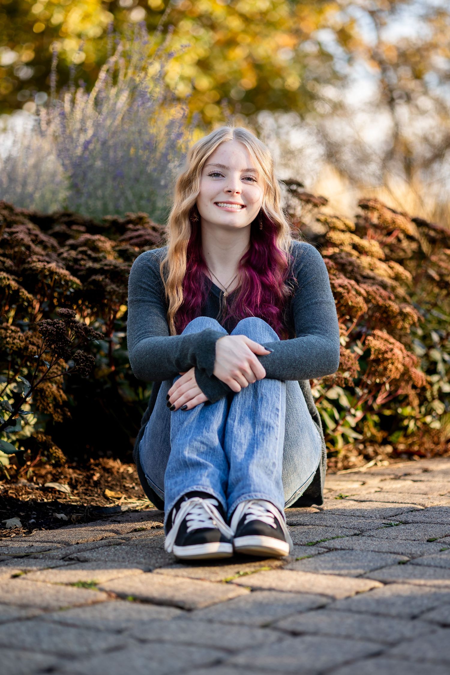 A person in a gray sweater and jeans sits on stone pavement with autumn foliage in the background during golden hour.