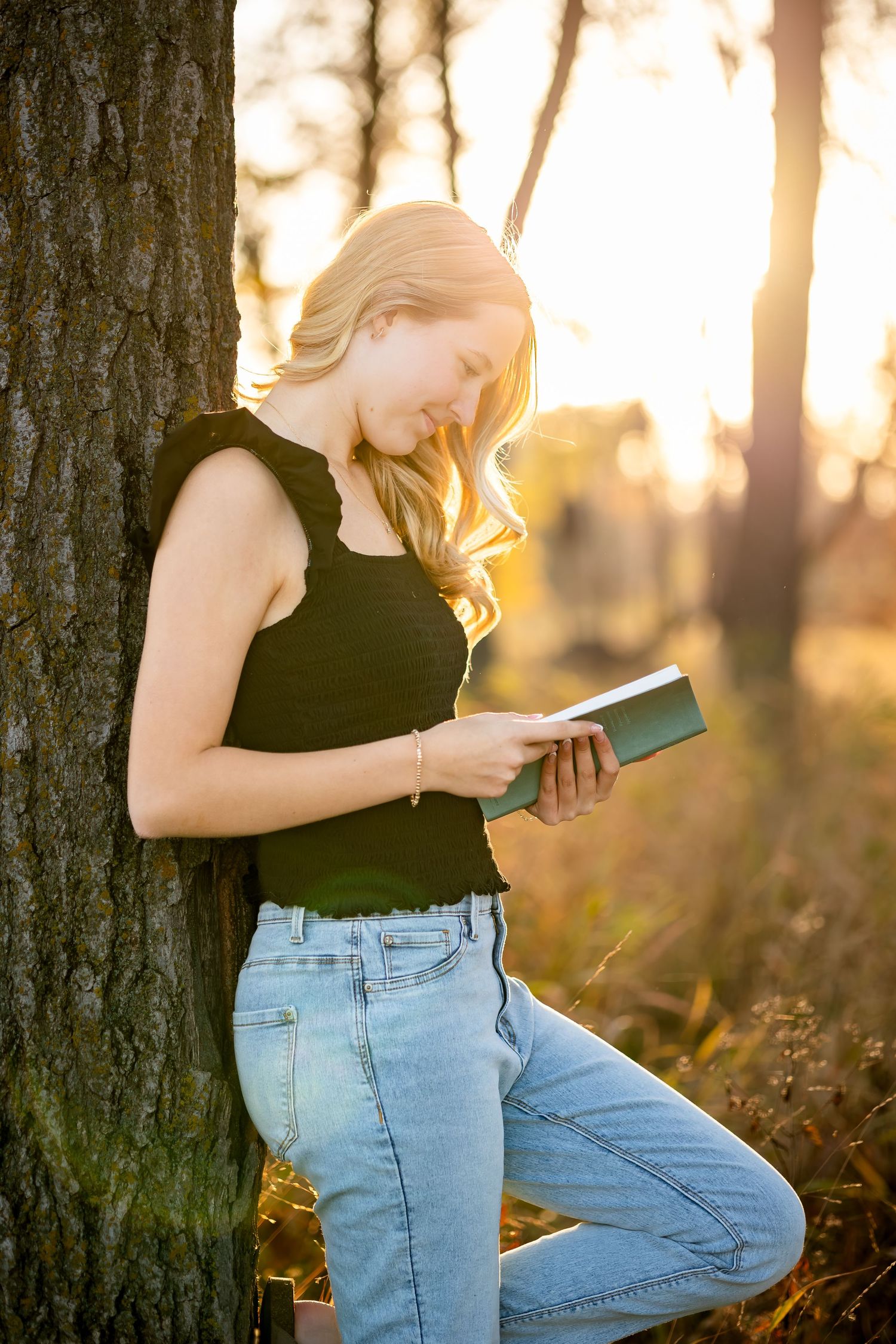 Person in black tank top and light blue jeans leans against tree trunk during golden sunset in nature setting.
