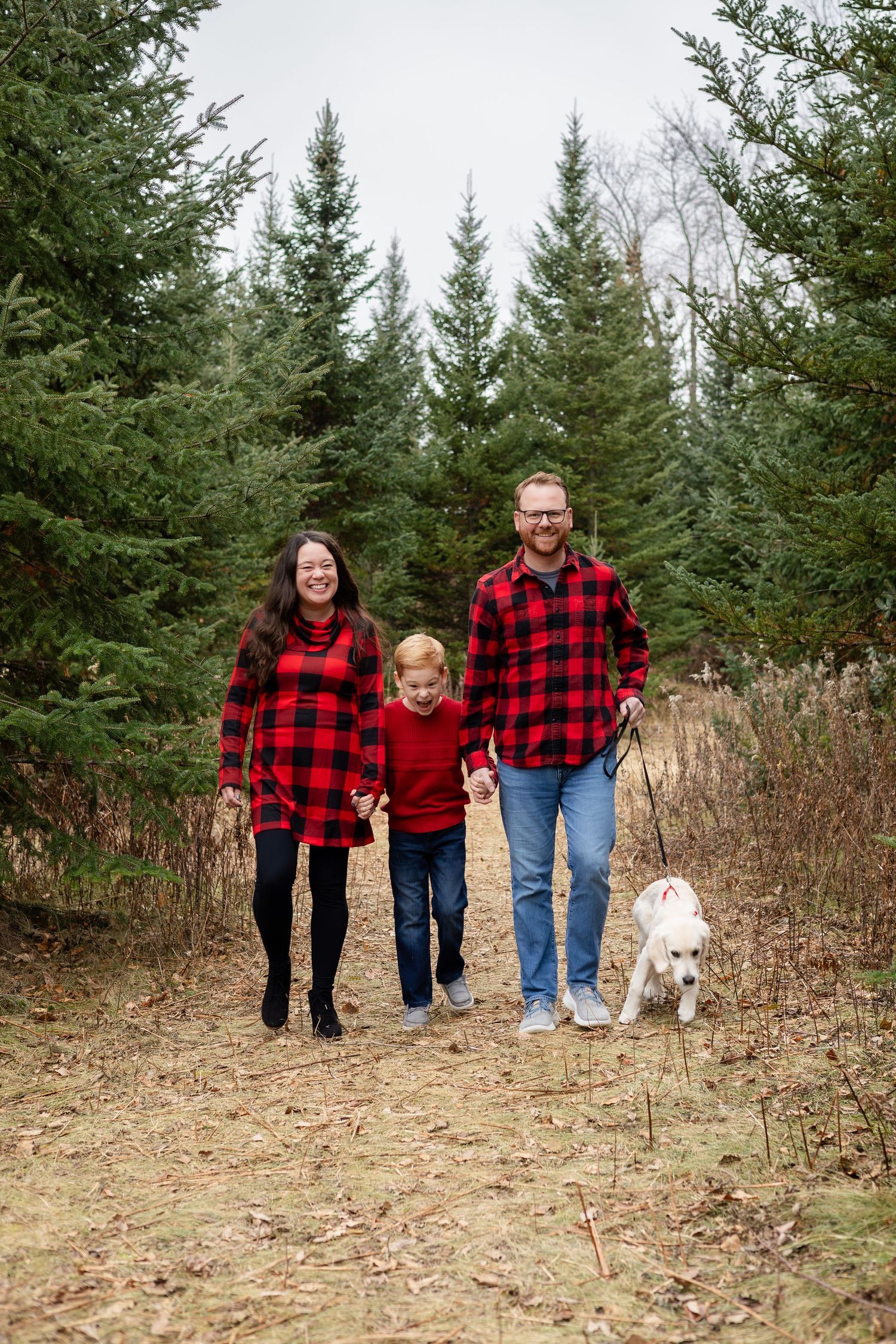 A family in matching buffalo plaid shirts walks together with their white dog through a forest clearing.