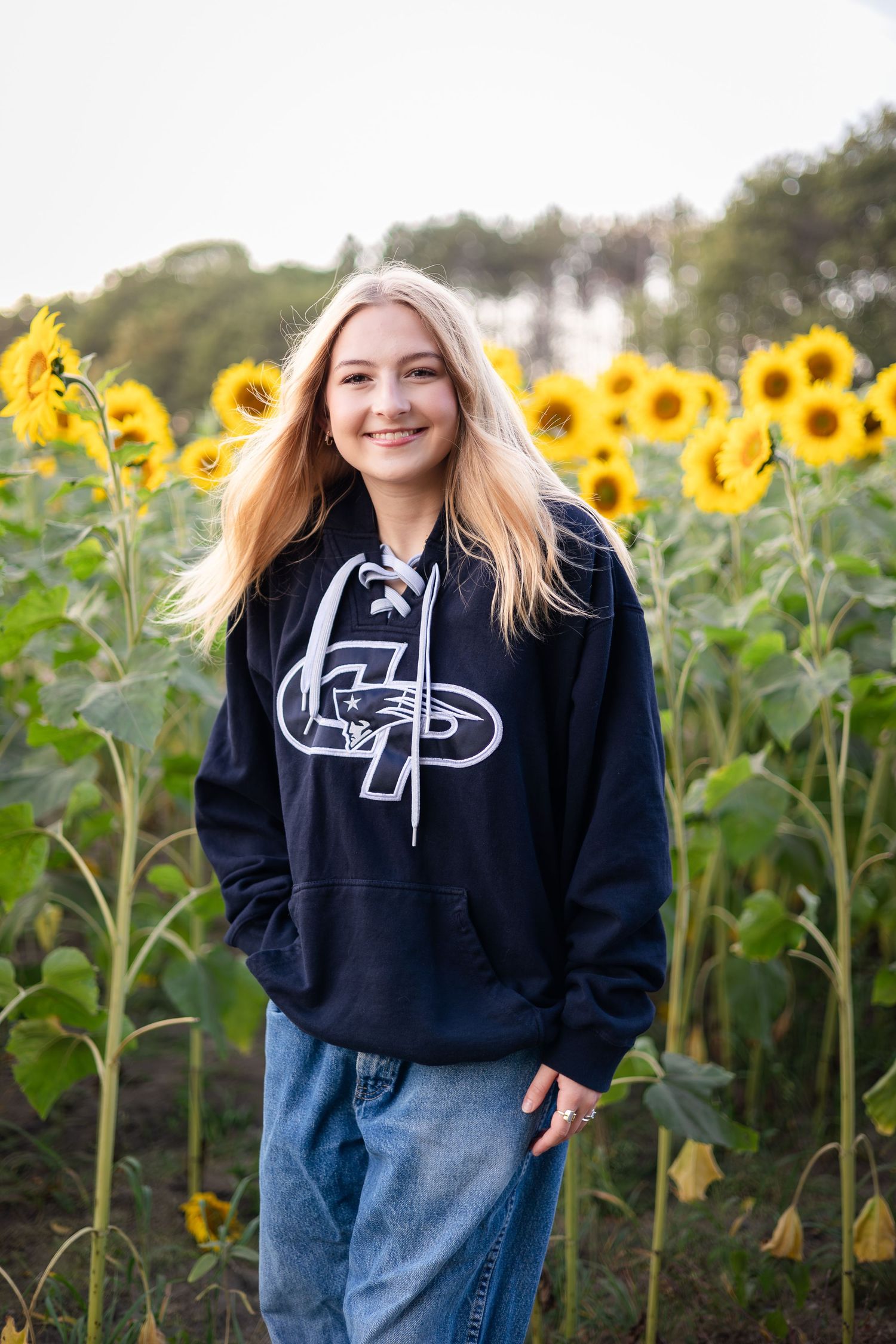 Woman in black hoodie and jeans poses cheerfully among blooming sunflowers.