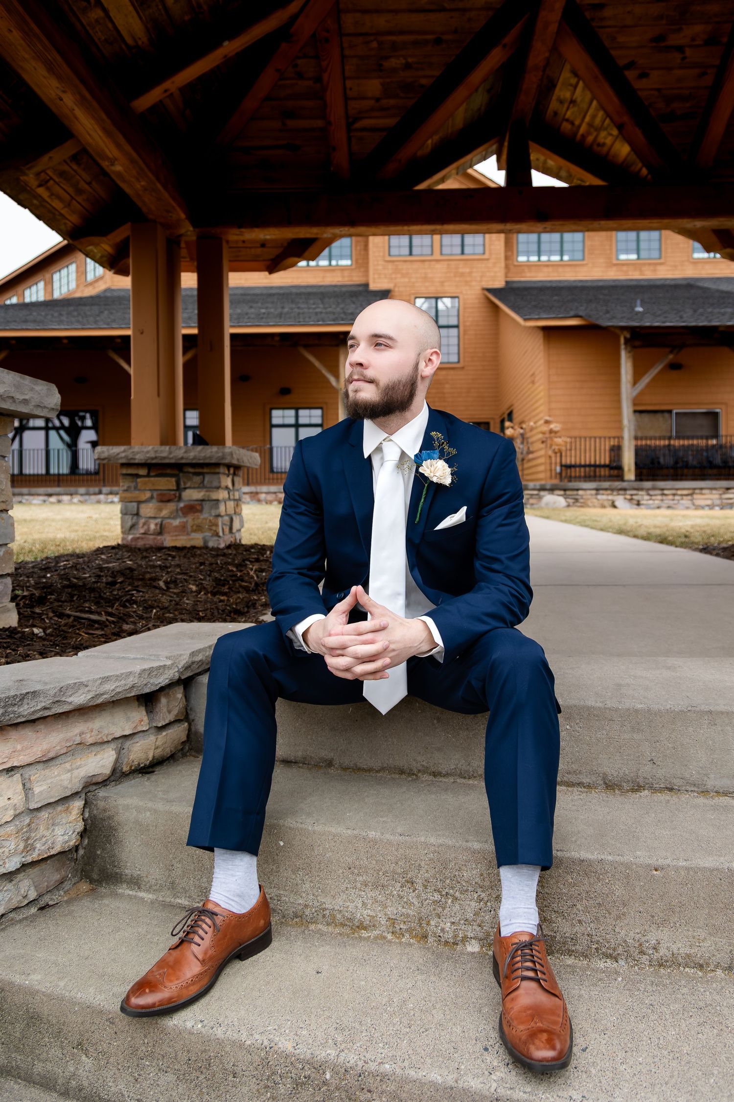 Groom in navy suit with boutonniere sits on stone steps outside rustic wedding venue.