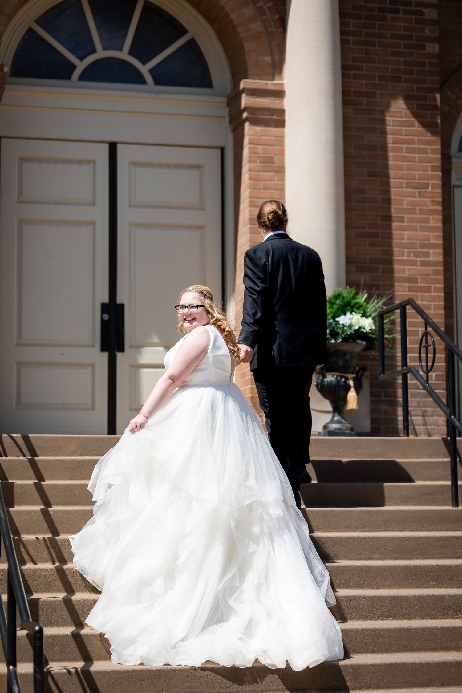 Bride in ruffled white ball gown walks up church steps holding grooms hand as he leads her inside.