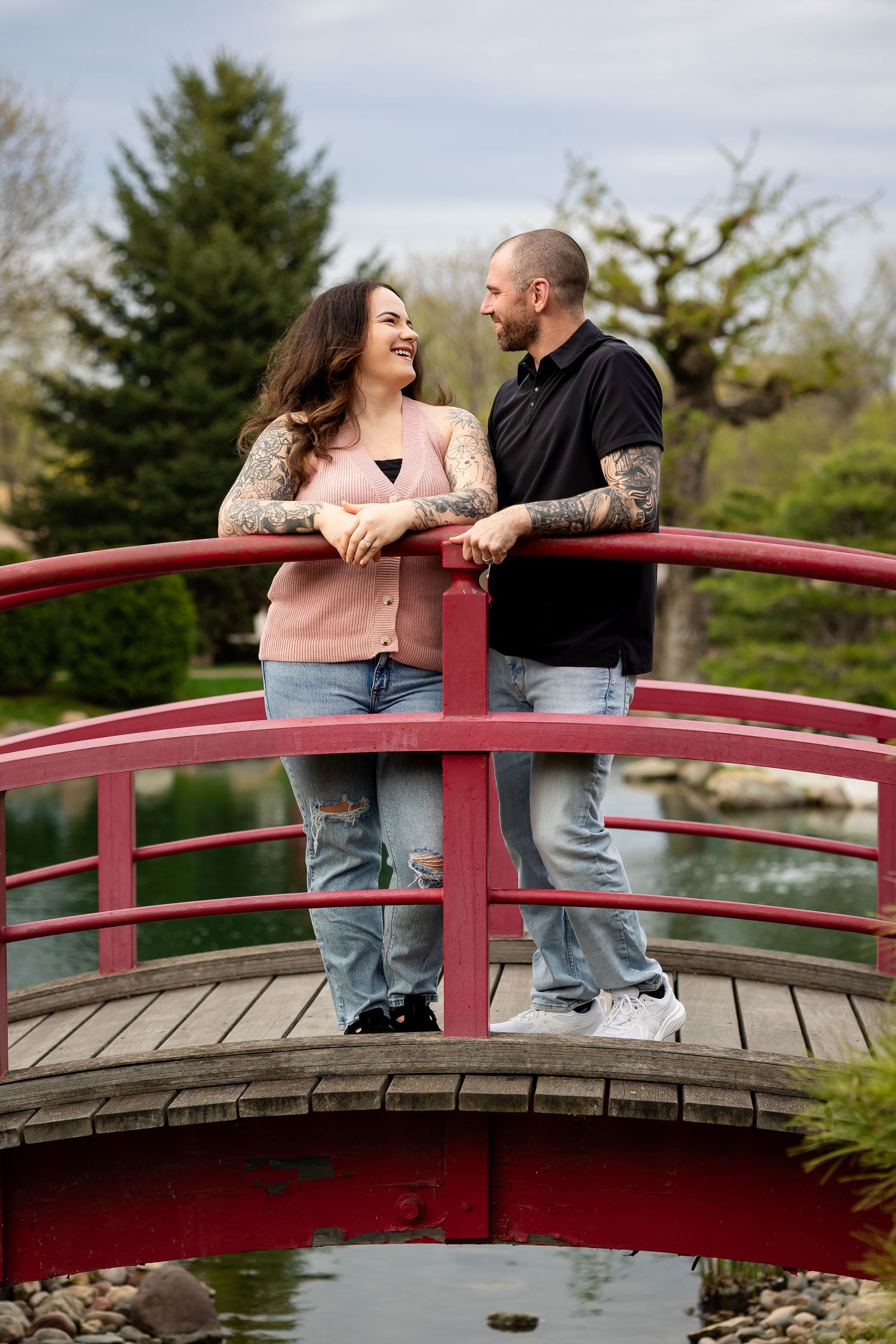 Couple with tattoos smiling at each other on a red arched bridge over a pond in a garden park.