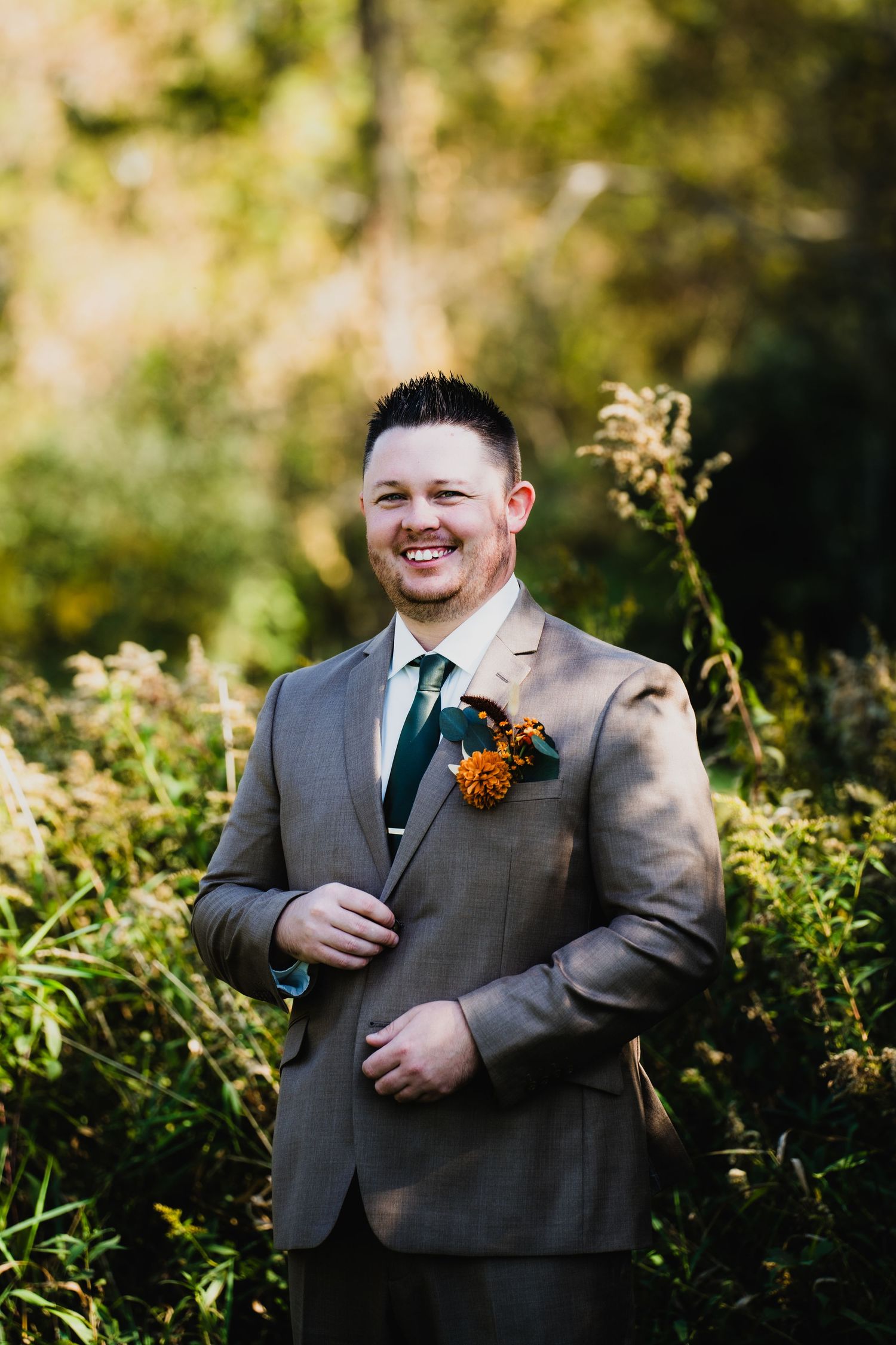Groom in gray suit with orange boutonniere poses in natural outdoor setting.