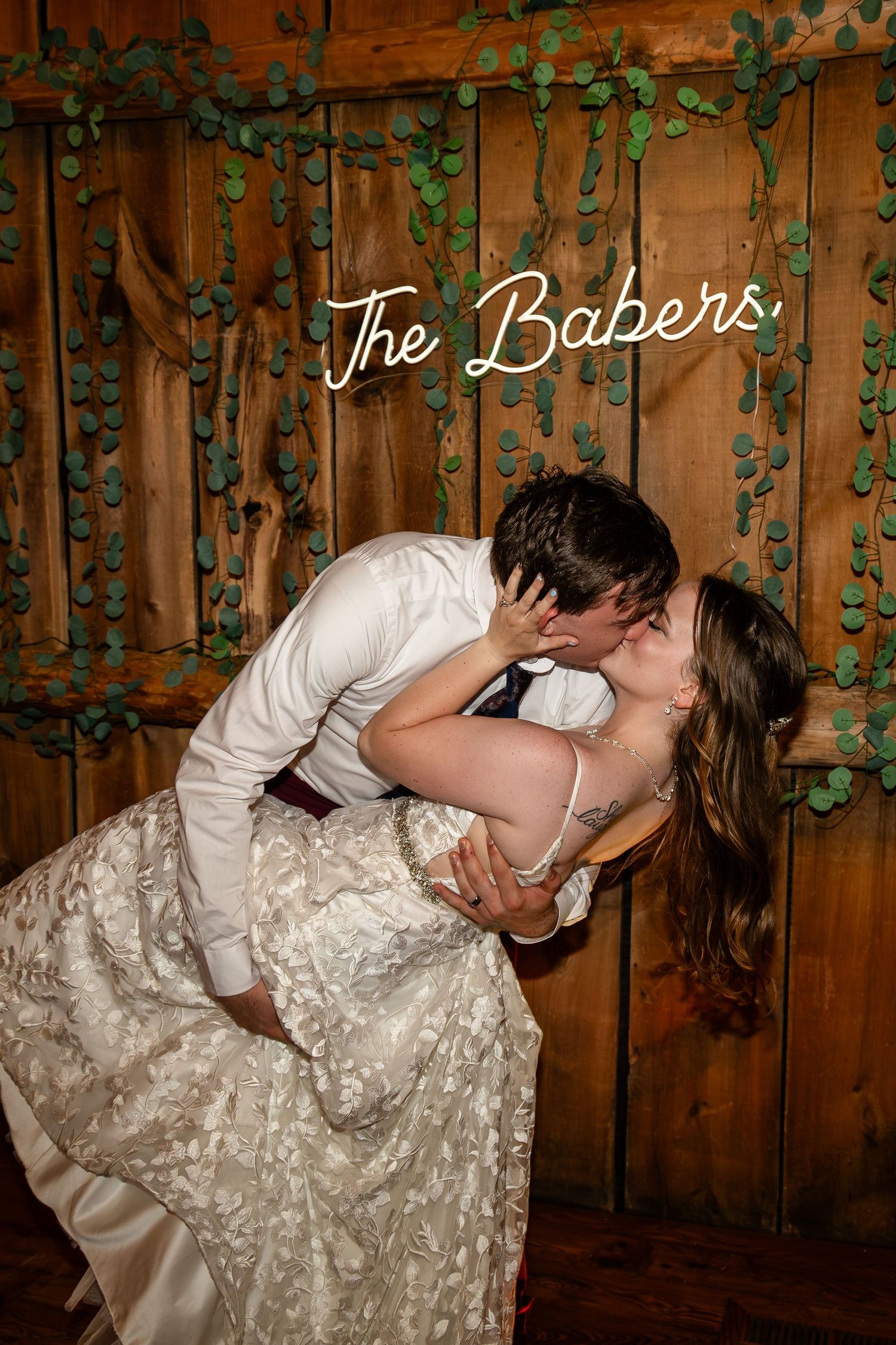 A wedding couple shares a romantic dip kiss against a rustic wooden backdrop with ivy.