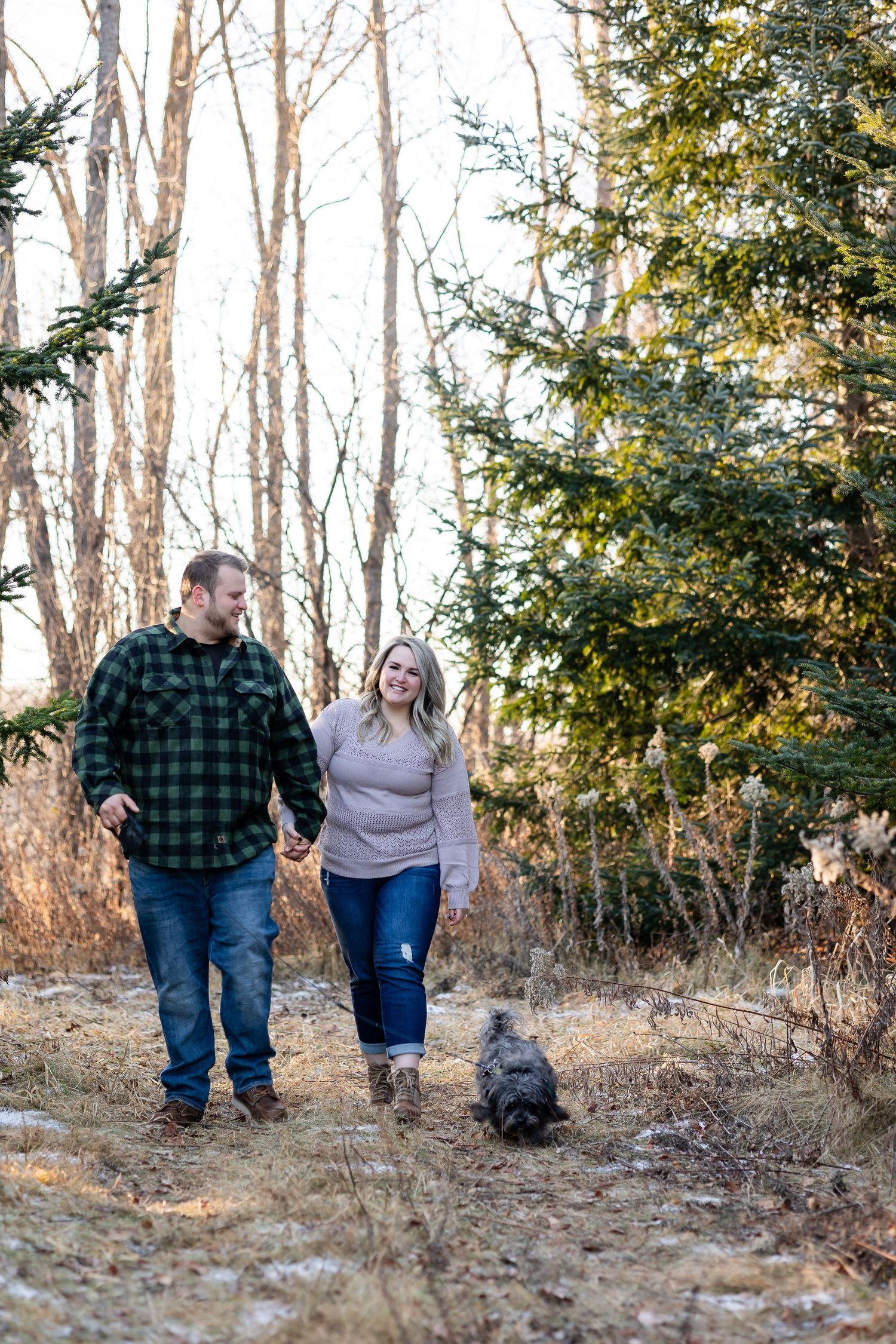A couple walking with their black dog through a wooded area during a winter photo session.