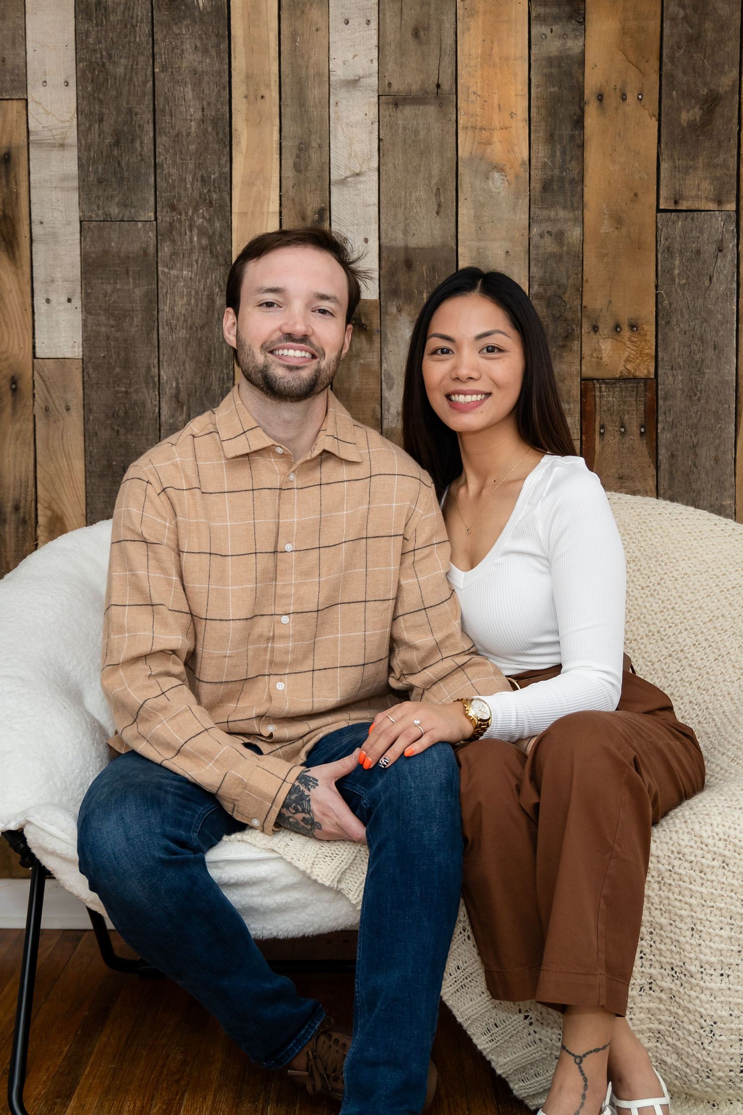 A couple sits together on a white couch against a rustic wooden wall background.