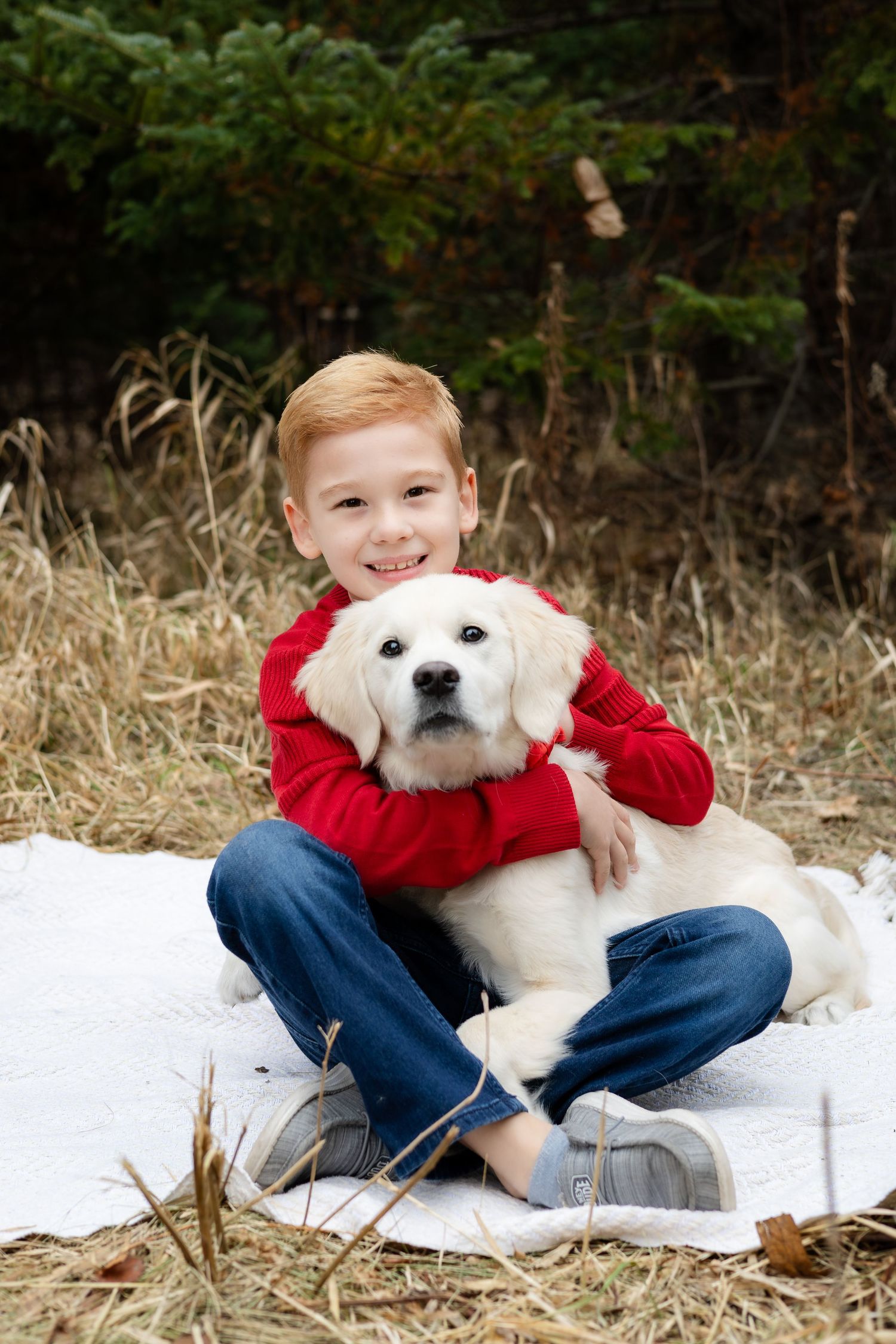 A child in a red sweater hugs a white dog while sitting on a blanket outdoors.