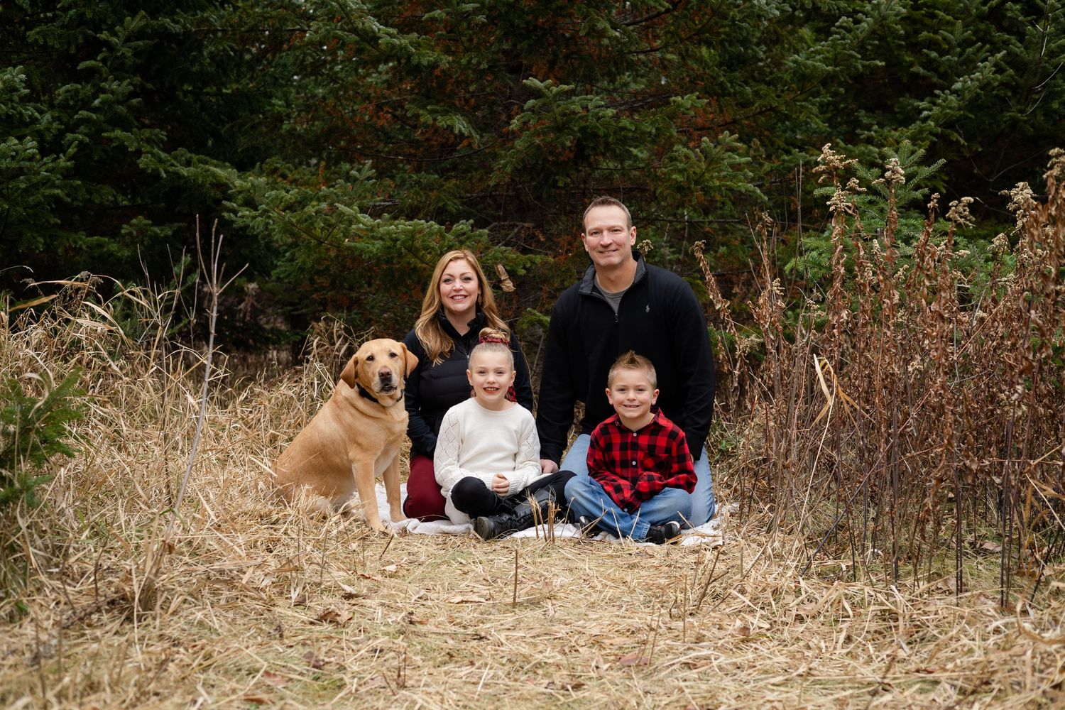 Family and dog relaxing in a natural outdoor setting with evergreen trees and dried grass.