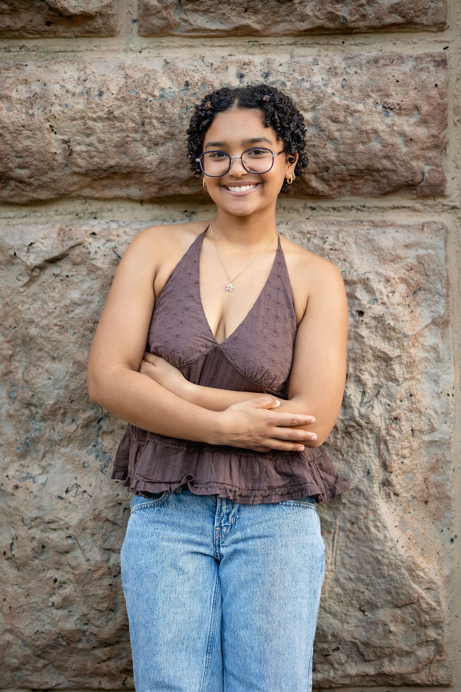 A person in a brown tank top and light jeans poses casually against a textured stone wall.