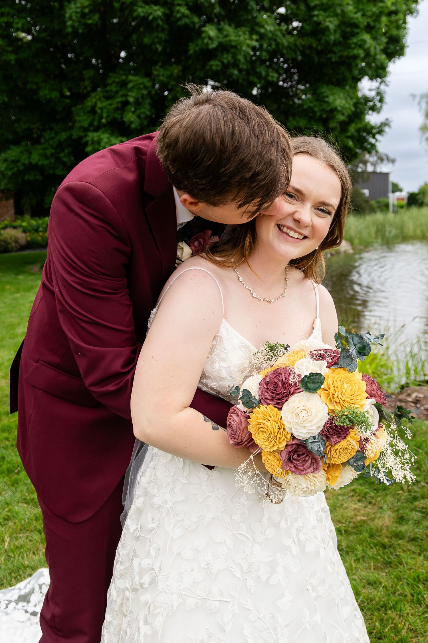 A romantic wedding couple embraces outdoors near a lake with a vibrant autumn-colored bouquet of roses and flowers.