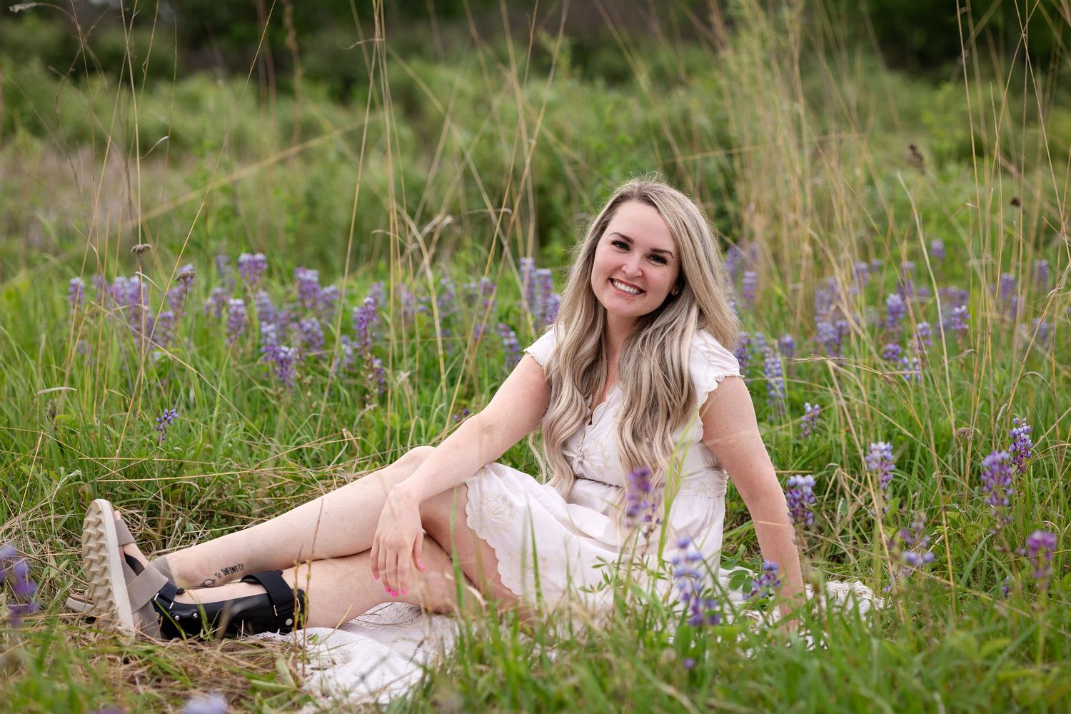 Smiling blonde woman in white dress sitting in a wildflower field with purple lupines and green grass.