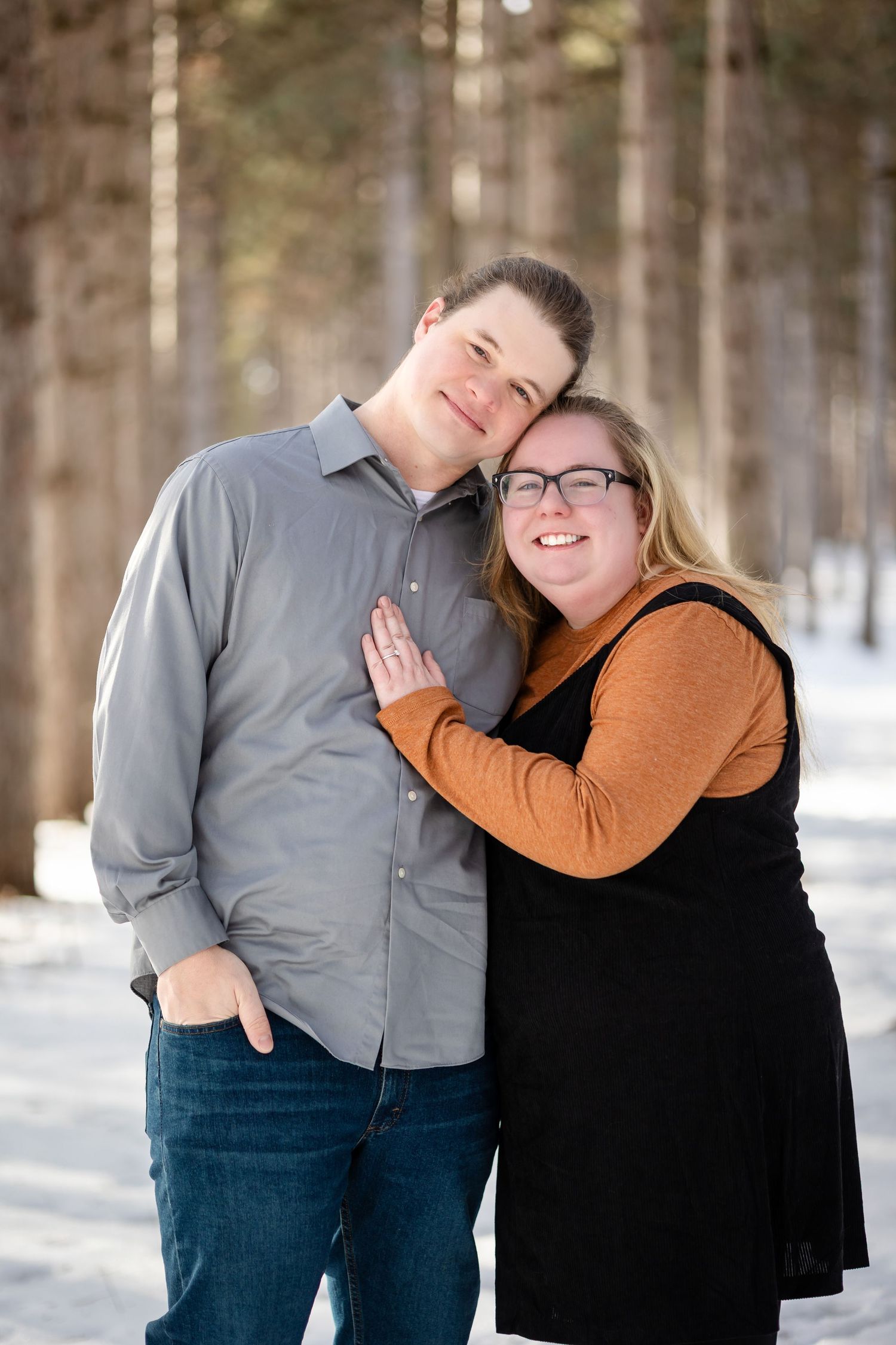 Couple embracing and smiling in snowy winter forest, woman in orange sweater and black dress leaning on man in gray shirt.