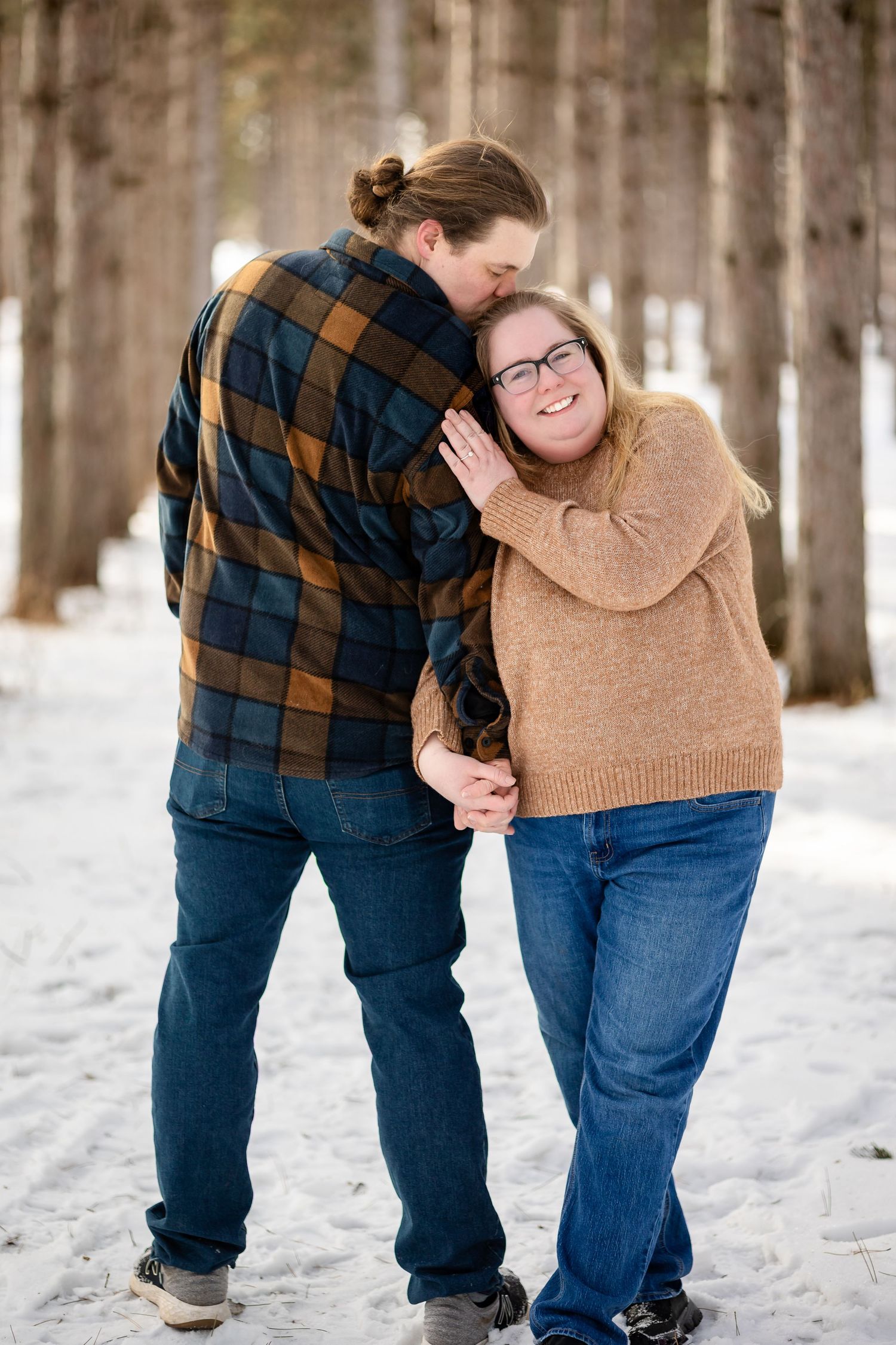 Man kissing woman on the head while holding hands in a snowy pine tree lined path in winter.