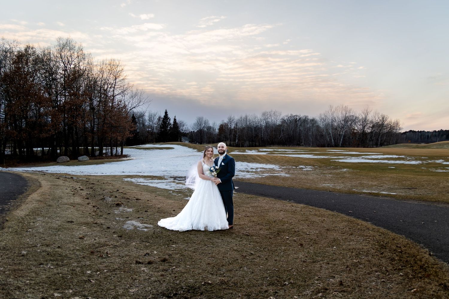 Bride and groom share a romantic moment on a winding path surrounded by a winter landscape at sunset.