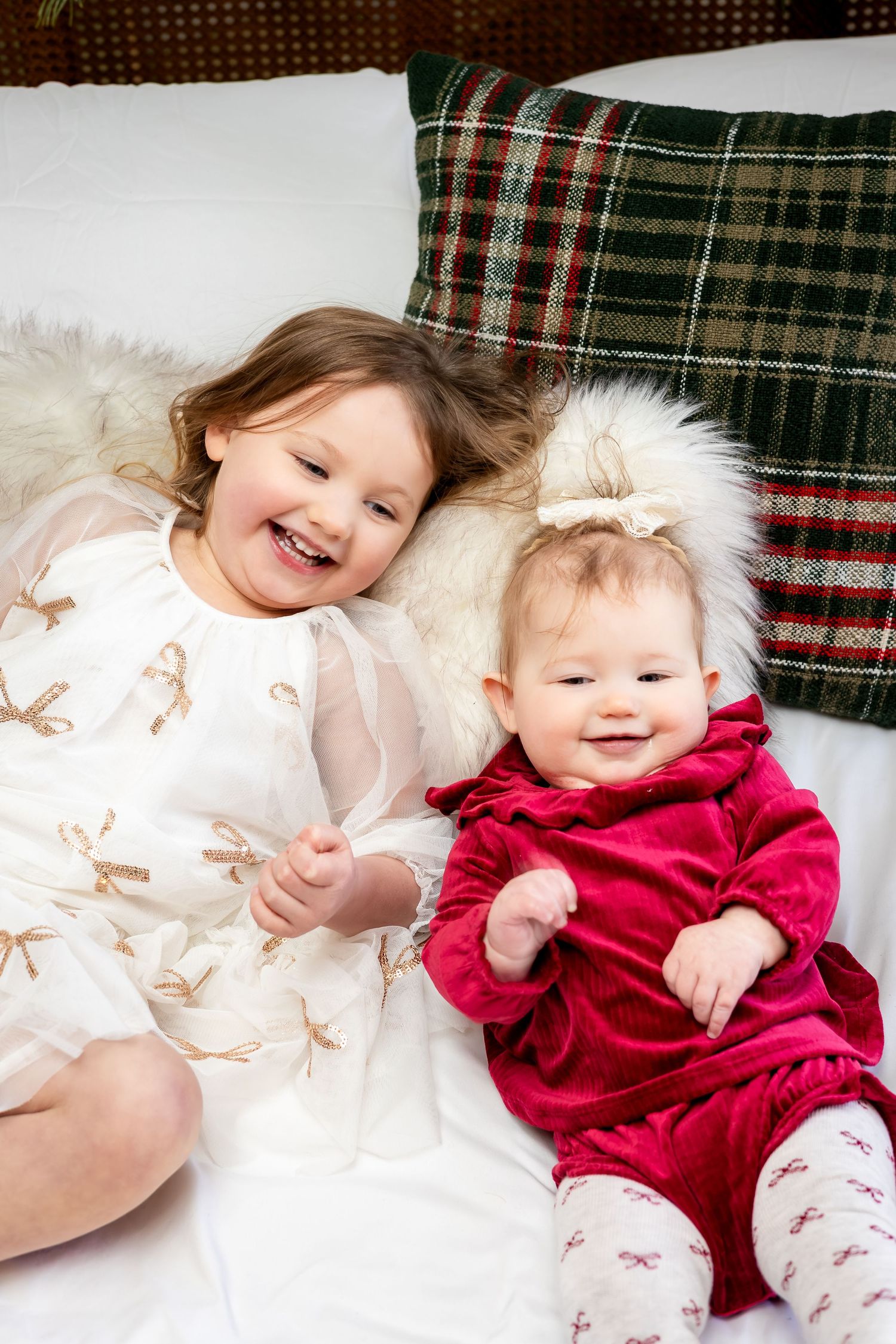 Two cheerful young sisters in holiday dresses laugh together on a white bed with plaid Christmas pillows.