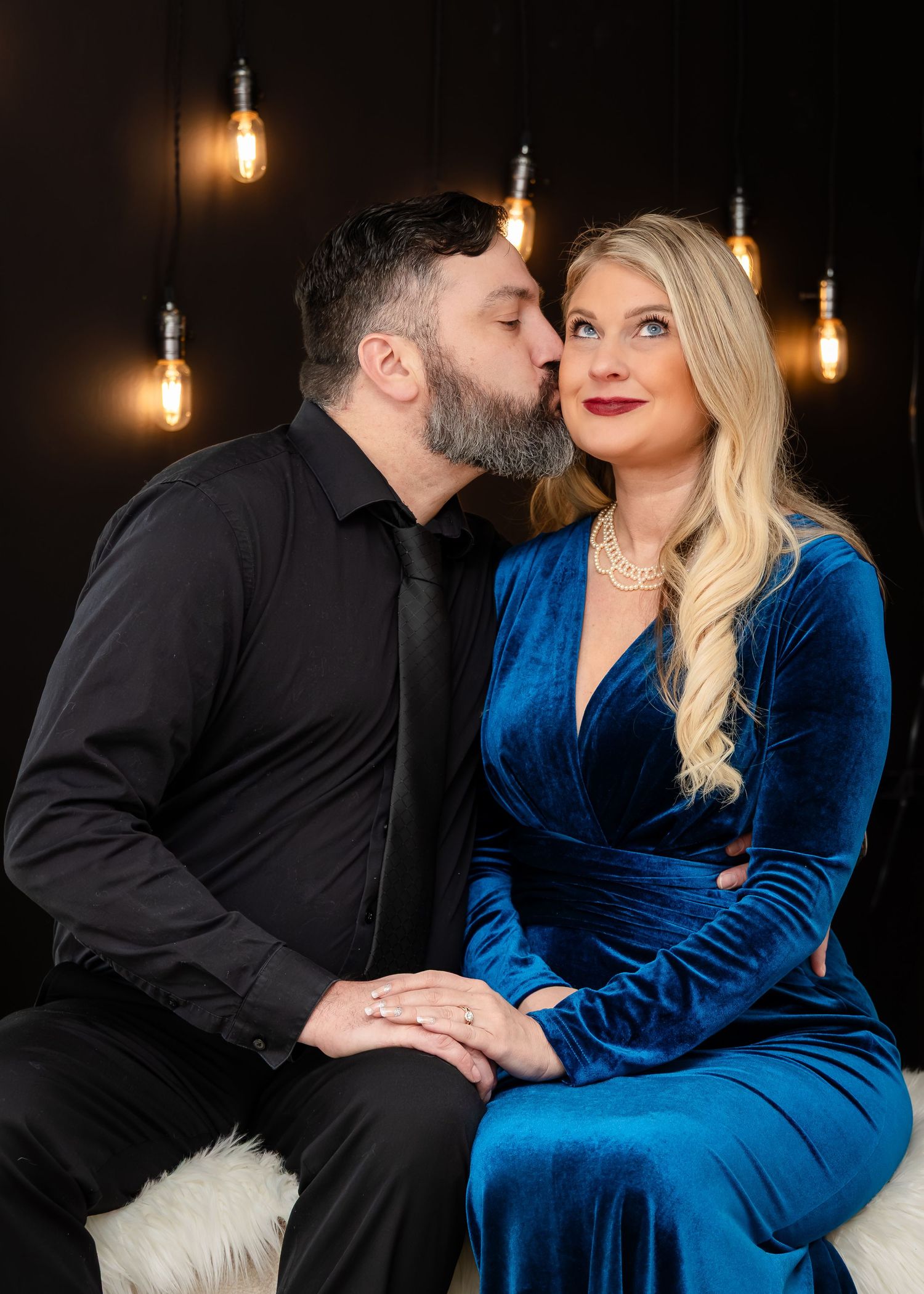 Couple in elegant attire, man in black shirt kissing woman in blue velvet dress, romantic portrait with Edison bulb backdrop.