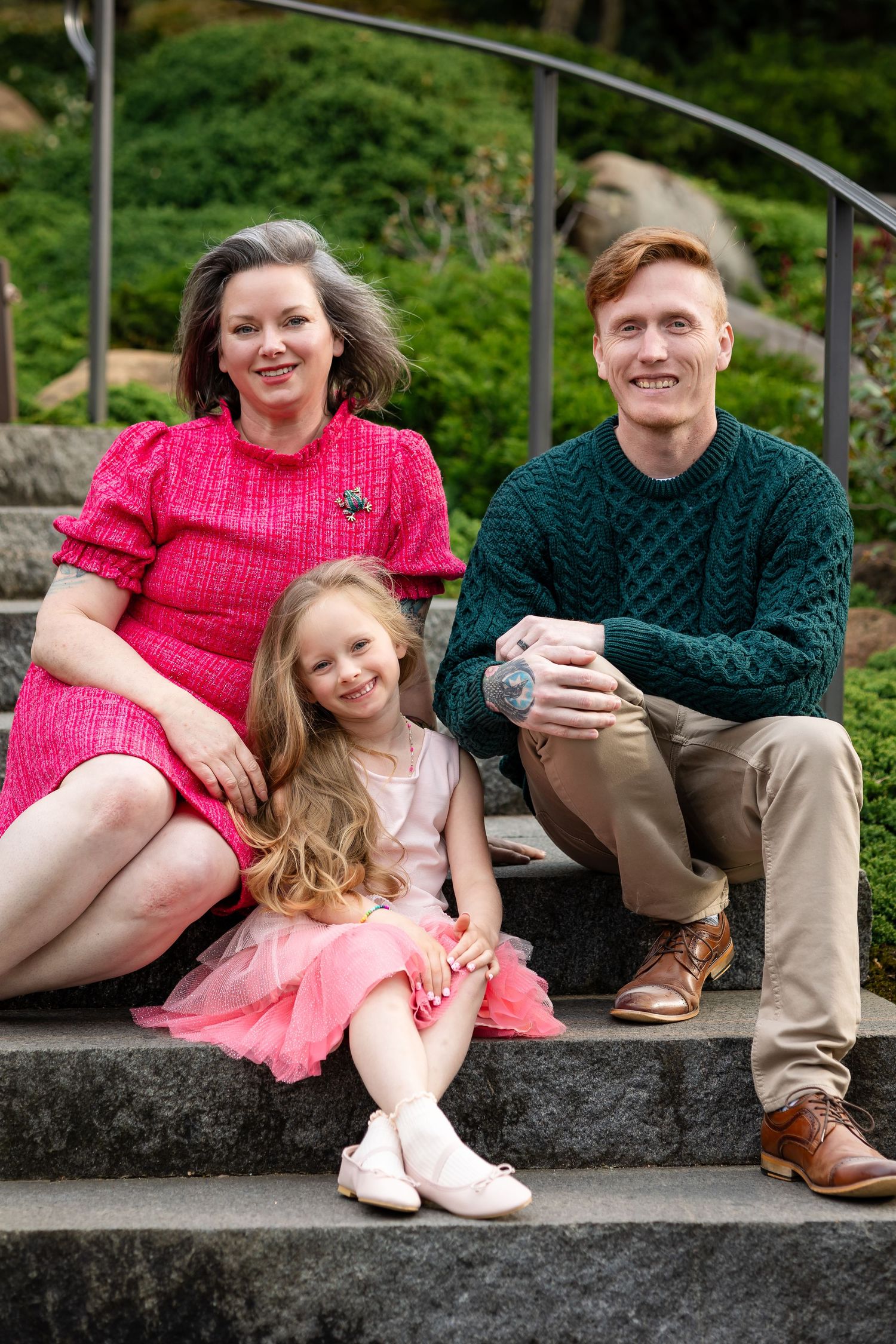 Family of three smiling on outdoor stone steps, woman in pink dress, man in green sweater, young girl in pink tutu.