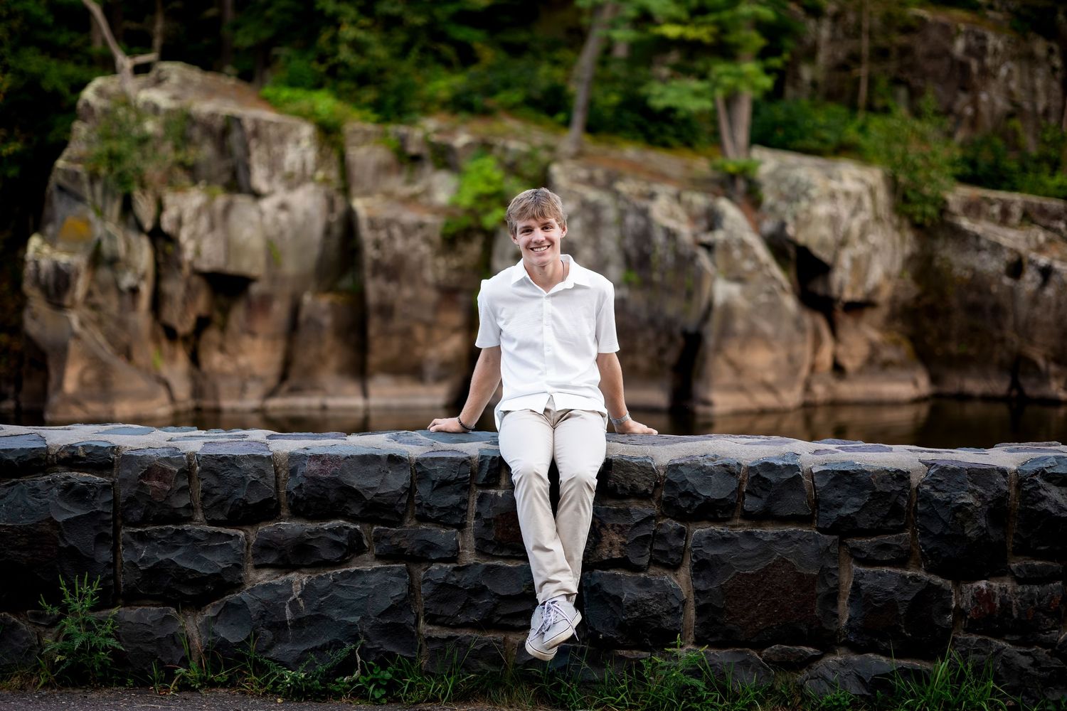 Person in white outfit sits casually on stone wall with rocky cliffs in background.