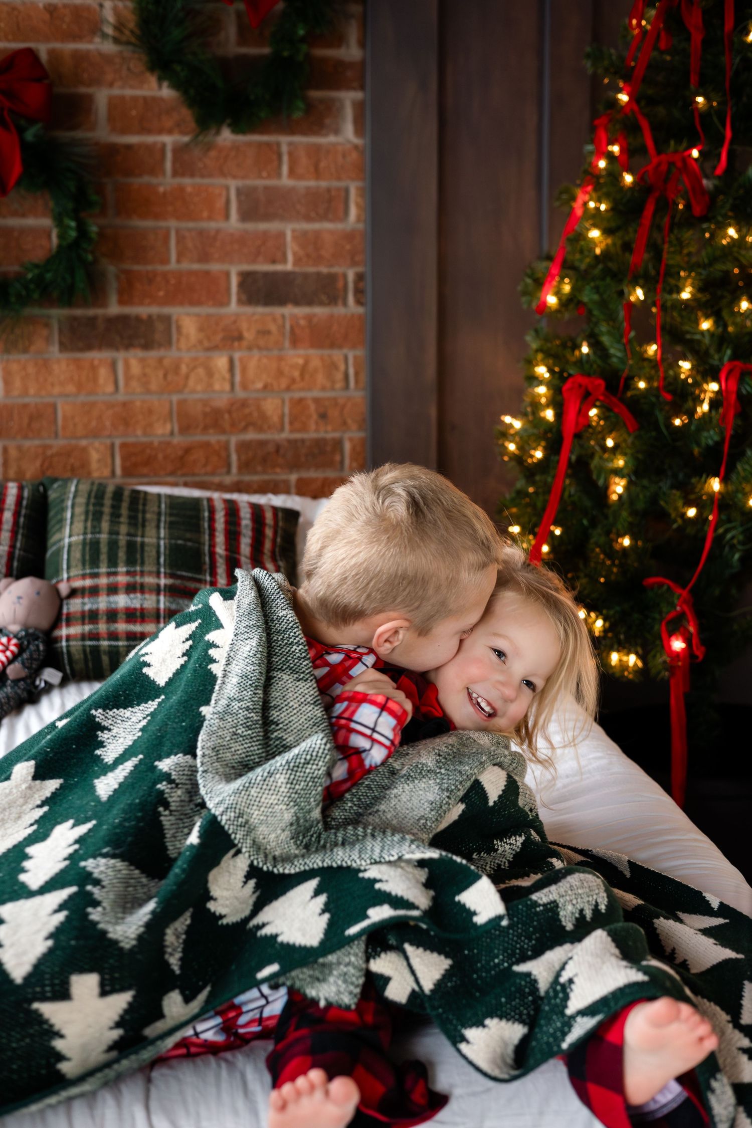 Children snuggling under festive green and white tree patterned blanket by Christmas decorations.