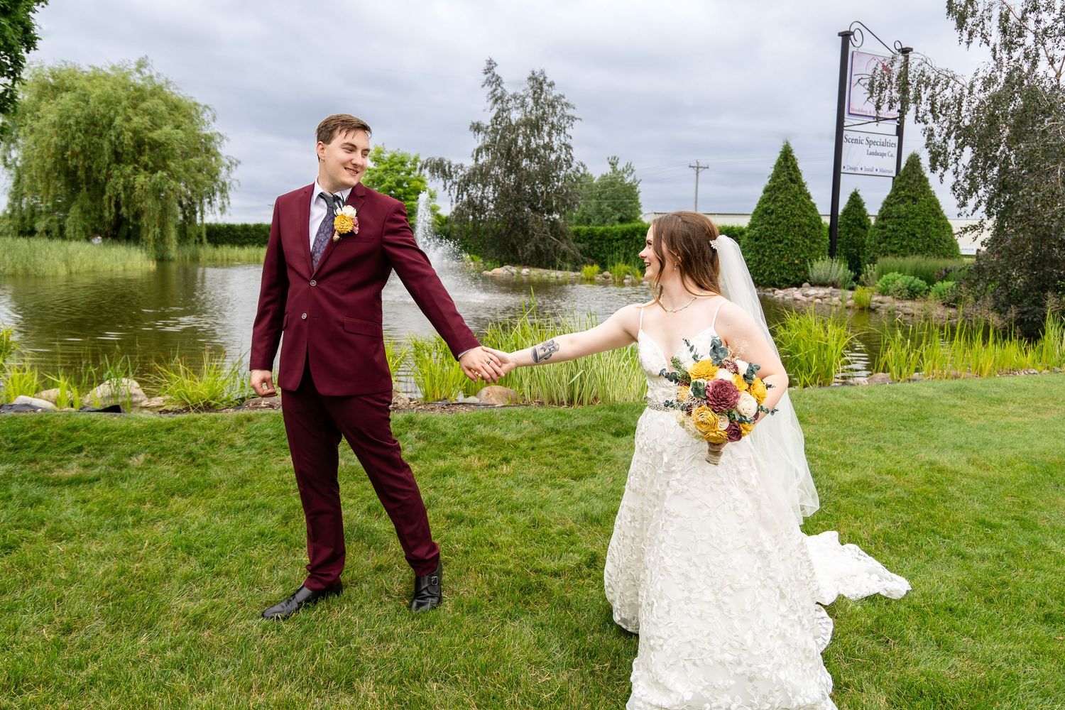Wedding couple dances playfully on green lawn in burgundy suit and white gown.