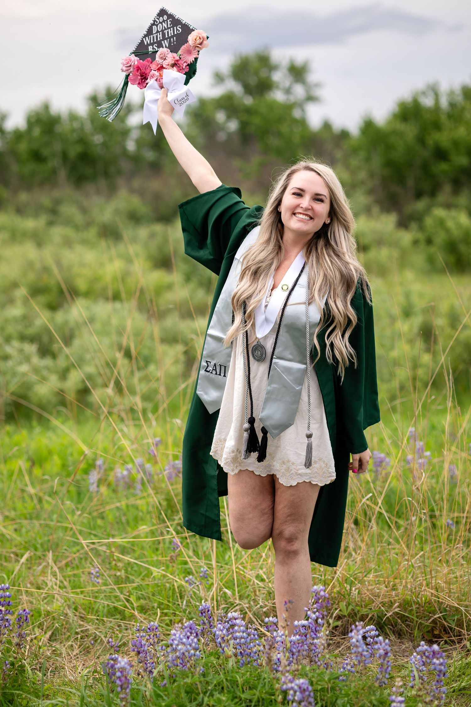 Smiling graduate in green gown holds decorated cap in wildflower field with purple lupines.