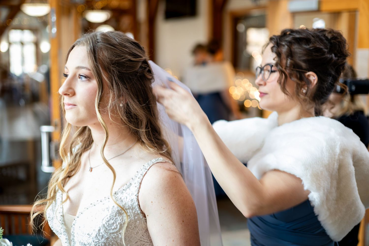 Bridesmaid carefully adjusts the brides veil inside a warmly lit rustic wedding venue before the ceremony.